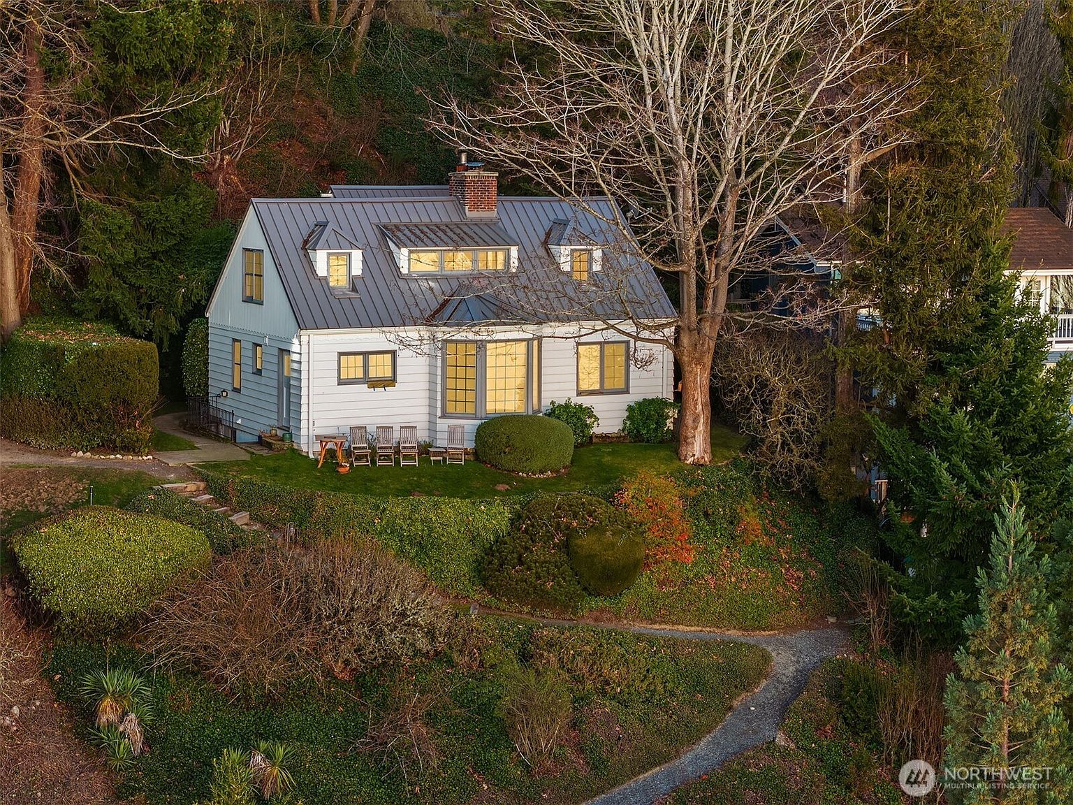 This elevated, slightly angled aerial view captures a charming, white-sided cottage nestled into a lush, sloped landscape. The home features a distinctive metal roof, multiple dormer windows, and a cozy backyard patio area with outdoor seating, all surrounded by mature trees and dense greenery. The perspective emphasizes the home's secluded, natural setting and its integration into the hillside terrain.