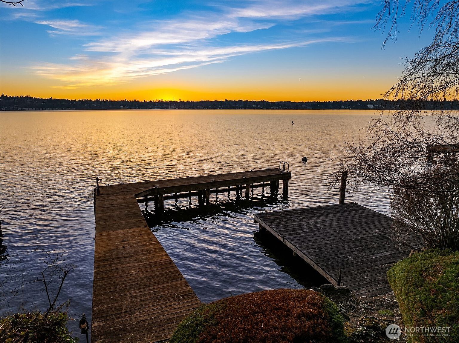 This serene waterfront view captures a wooden dock extending into a calm lake during a vibrant sunset. The composition highlights the peaceful outdoor living space, featuring a multi-level dock structure set against a backdrop of a distant treeline and a glowing horizon. The perspective is from the shoreline, emphasizing the direct water access and the tranquil atmosphere of the property.