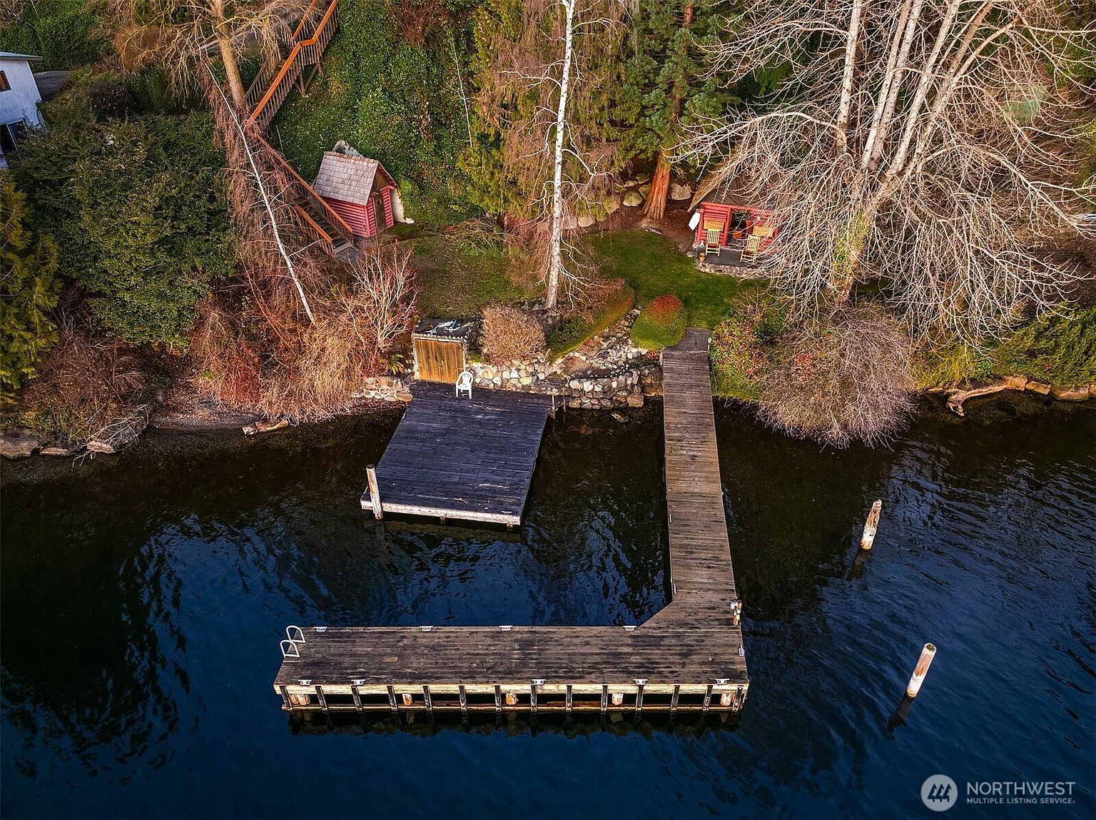 This high-angle aerial shot captures a serene waterfront property featuring a large, L-shaped wooden dock extending into a calm lake. The shoreline is landscaped with a small red cabin, a secondary shed, and lush greenery, all connected by a wooden staircase leading up the hillside. The perspective emphasizes the tranquil, private nature of the retreat, highlighting the seamless integration between the rustic structures and the natural lakeside environment.