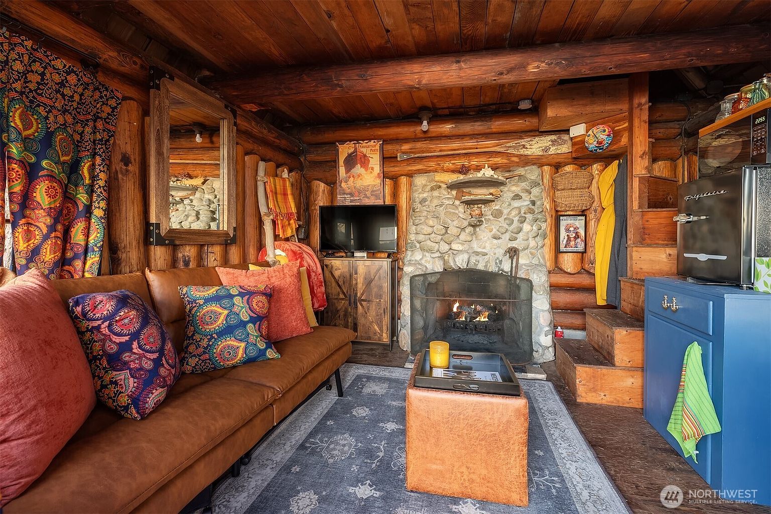 This cozy, rustic living room features log cabin walls, a stone fireplace with a crackling fire, and a warm, inviting atmosphere. The space is furnished with a brown leather sofa adorned with colorful patterned pillows, a matching ottoman, and a blue area rug, all set against a backdrop of eclectic decor including a vintage-style poster and a wooden oar. The perspective is a wide-angle shot from the entrance, capturing the intimate, cabin-like charm and the transition to a small kitchen area with stairs.