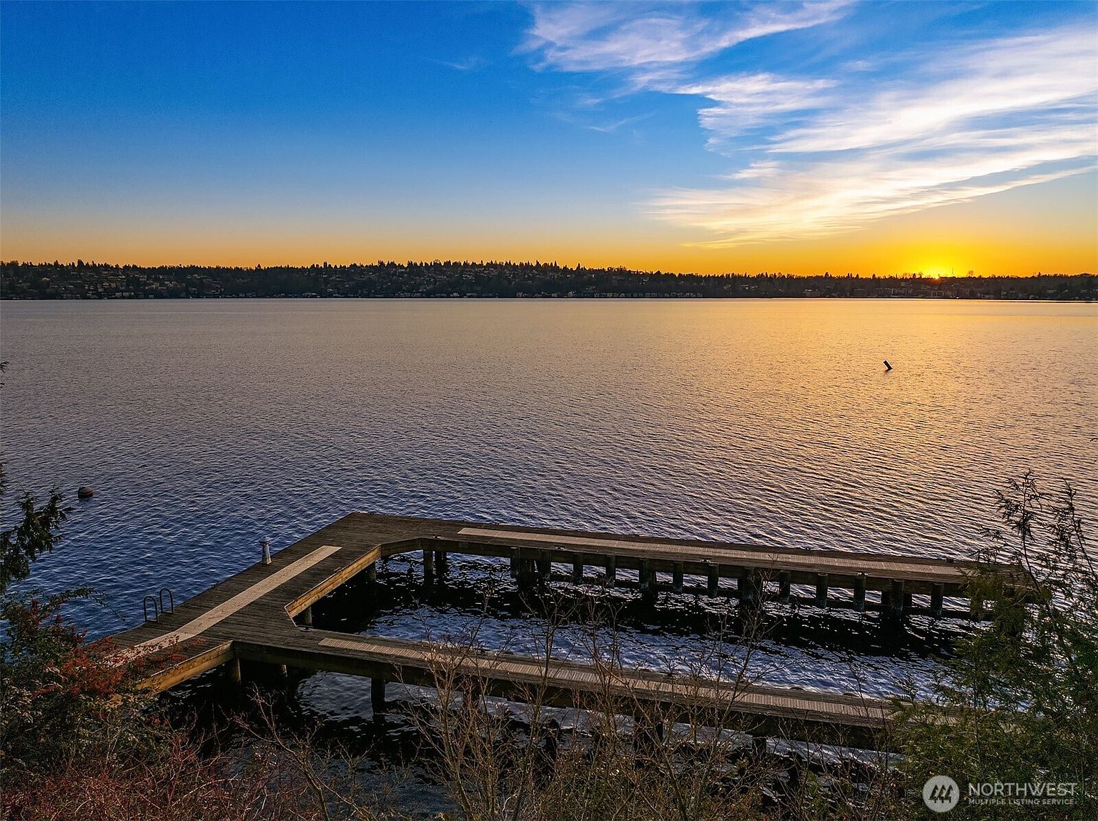 This elevated perspective captures a serene waterfront view at sunset, featuring a wooden dock extending into a calm lake. The horizon is lined with a distant treeline silhouetted against a vibrant, gradient sky transitioning from deep blue to warm orange. The scene evokes a peaceful, high-end lifestyle, highlighting the property's direct water access and scenic natural surroundings.