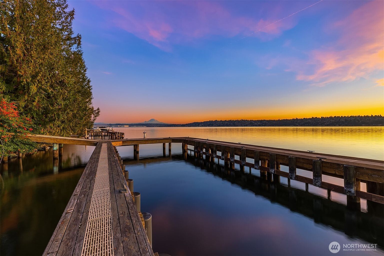 This scenic waterfront view features a long, weathered wooden dock extending into a calm lake at sunset, with a distant snow-capped mountain visible on the horizon. The composition highlights the tranquil transition from the lush, tree-lined shoreline to the expansive, colorful sky reflecting on the water. The perspective is from the start of the dock, drawing the viewer's eye toward the serene horizon, emphasizing the property's premium waterfront access.