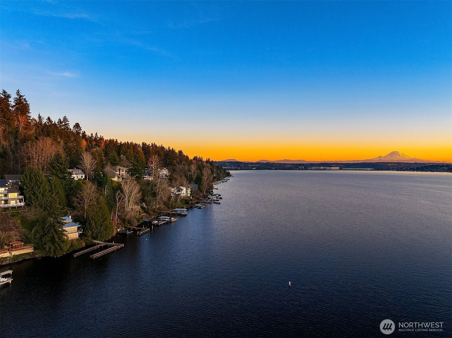 This stunning aerial view captures a picturesque waterfront property at sunset, showcasing a lush, tree-lined shoreline dotted with residential homes and private docks. The expansive, calm lake reflects the vibrant orange and blue hues of the twilight sky, with the majestic, snow-capped Mount Rainier visible in the distance. The perspective provides a serene and expansive look at the property's prime location and natural surroundings.