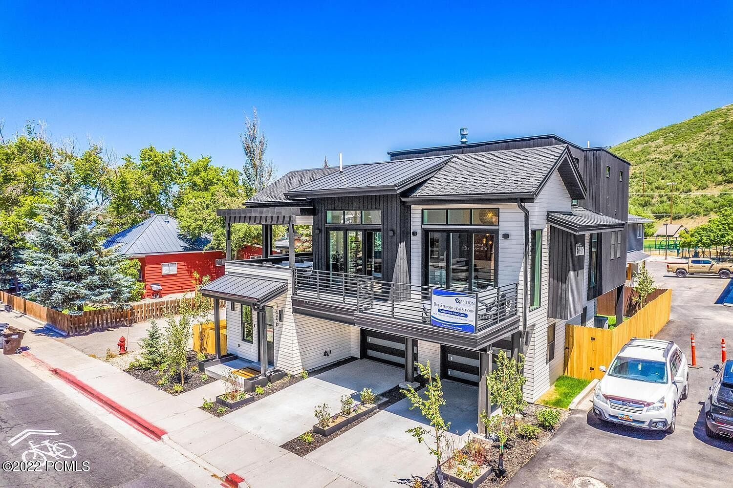 This is an eye-level, slightly elevated shot of a modern two-story home with a black and white exterior. The house features a balcony with metal railings, a covered patio, and a two-car garage. The landscaping is well-maintained with small trees and flower beds, and the overall impression is one of contemporary elegance and curb appeal.