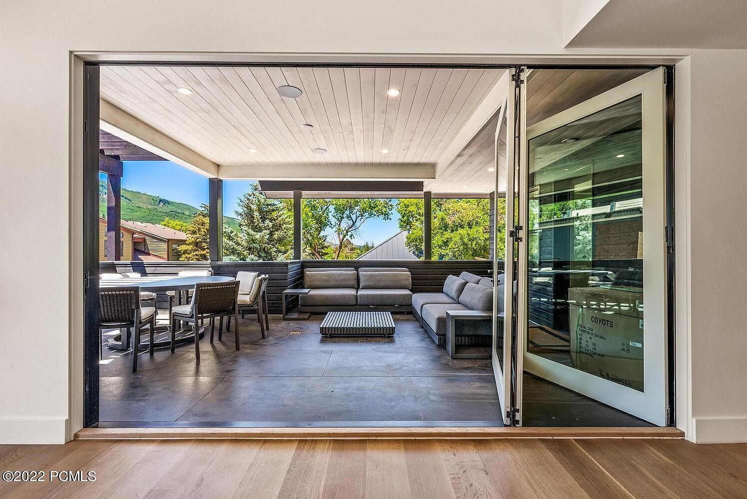 This image showcases a spacious outdoor patio or balcony area, seamlessly connected to the interior through large, folding glass doors. The space features a comfortable seating area with a gray sectional sofa and a modern coffee table, as well as a dining area with a round table and wicker chairs. The view from the patio includes lush greenery and a glimpse of the surrounding neighborhood, creating a relaxing and inviting atmosphere.