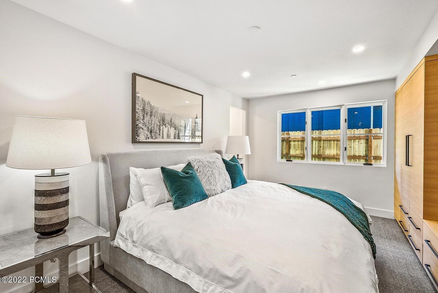 This is a bright and airy primary bedroom featuring a large bed with white linens, teal accent pillows, and a throw blanket. A framed landscape artwork hangs above the bed, and a modern lamp sits on a side table. Natural light streams in through the windows, creating a serene and inviting atmosphere.
