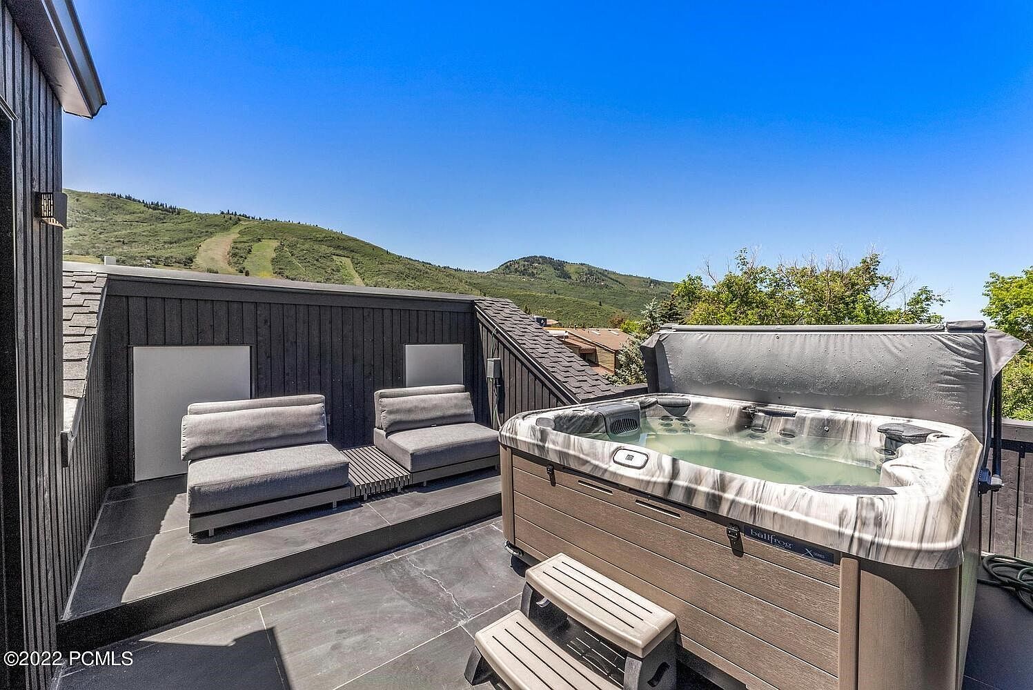 This image showcases a luxurious outdoor patio or deck area, complete with comfortable lounge seating and a hot tub. The deck is constructed with dark stone tiles, and the hot tub is surrounded by wood-like paneling and features a cover. The backdrop includes a scenic mountain view under a clear blue sky, enhancing the property's appeal for relaxation and entertainment.