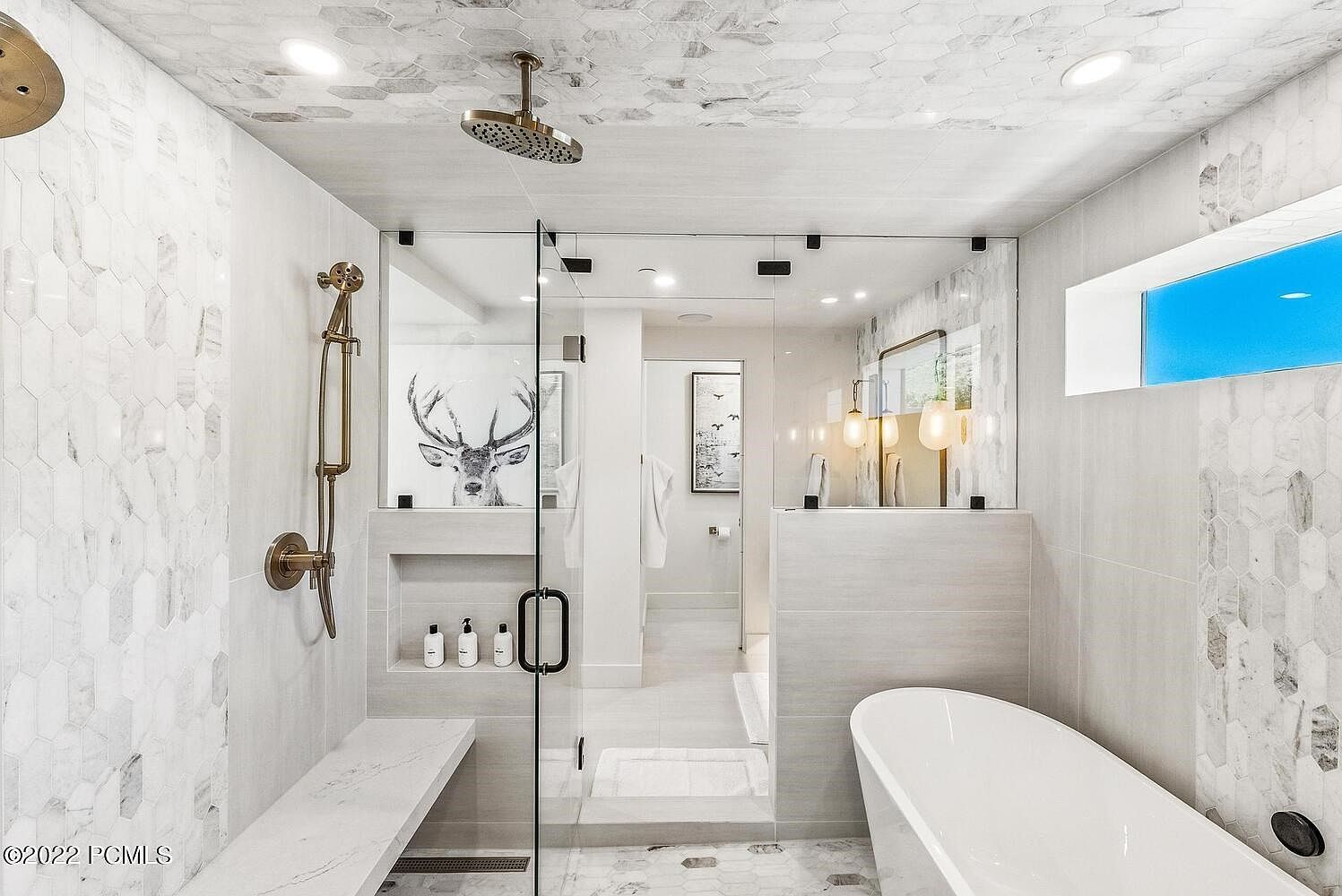 This is a luxurious primary bathroom featuring a glass-enclosed shower with a rainfall showerhead and a handheld sprayer. The walls and floor are adorned with elegant marble-like tiles in a hexagonal pattern, complemented by a built-in bench and niche for toiletries. A freestanding bathtub is visible on the right, and a window provides natural light, creating a spa-like atmosphere.