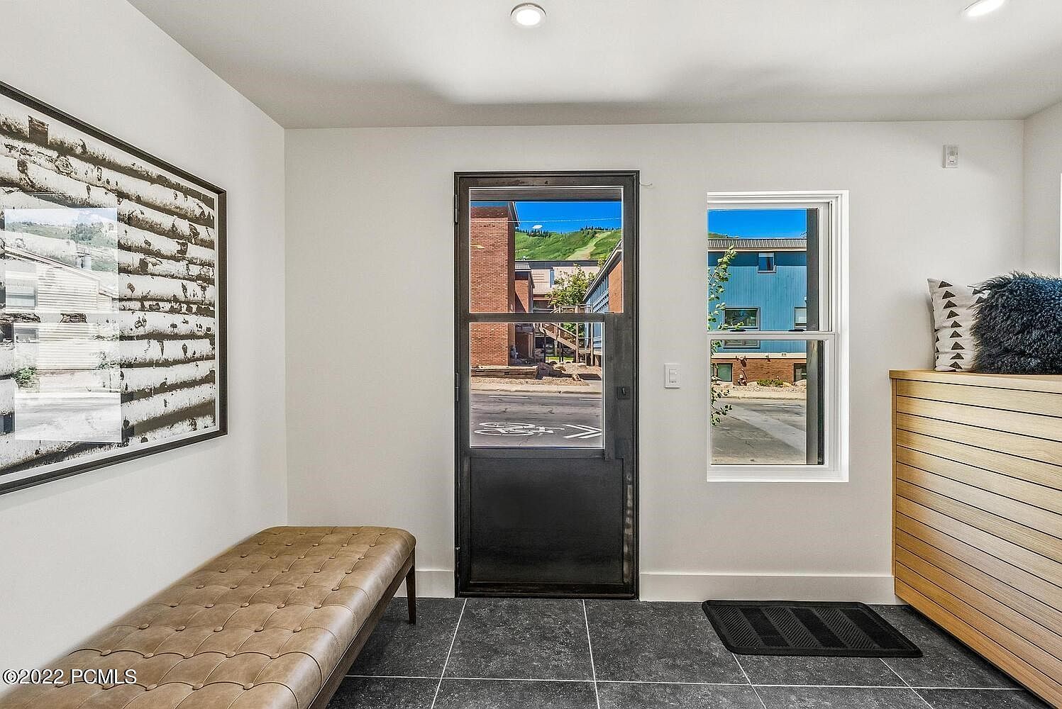 This interior shot showcases a modern entryway with a dark-framed glass door and a window providing natural light. A tufted bench sits against the wall, complemented by a contemporary art piece. The flooring is dark tile, and a wooden cabinet with horizontal slats adds a touch of warmth to the space.