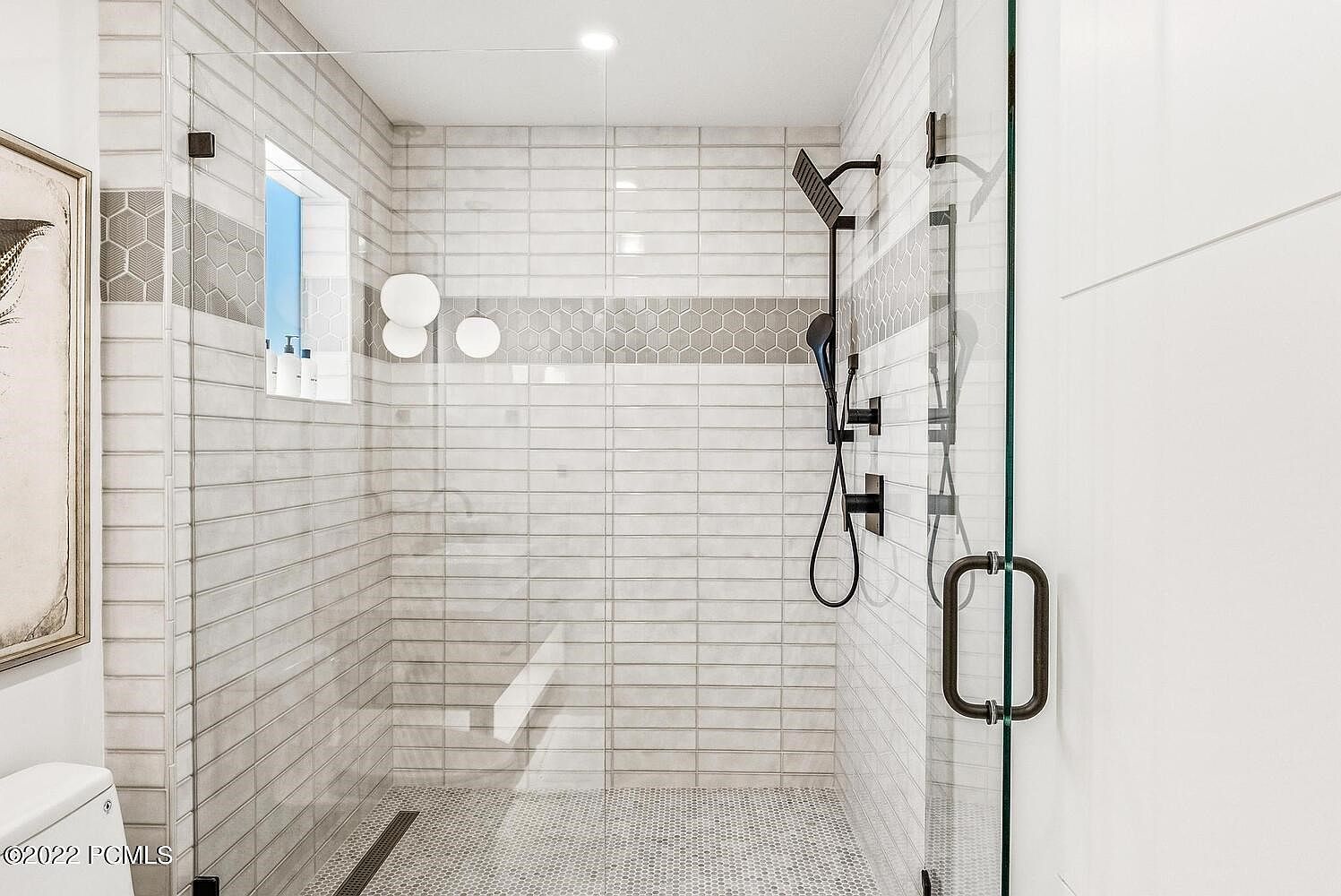 This is a well-lit primary bathroom featuring a modern shower with glass doors and a sleek, dark-toned shower fixture. The shower walls are tiled in a light gray horizontal pattern with a decorative band of hexagonal tiles. The floor is covered in small hexagonal tiles, and a linear drain is visible, adding to the contemporary design.