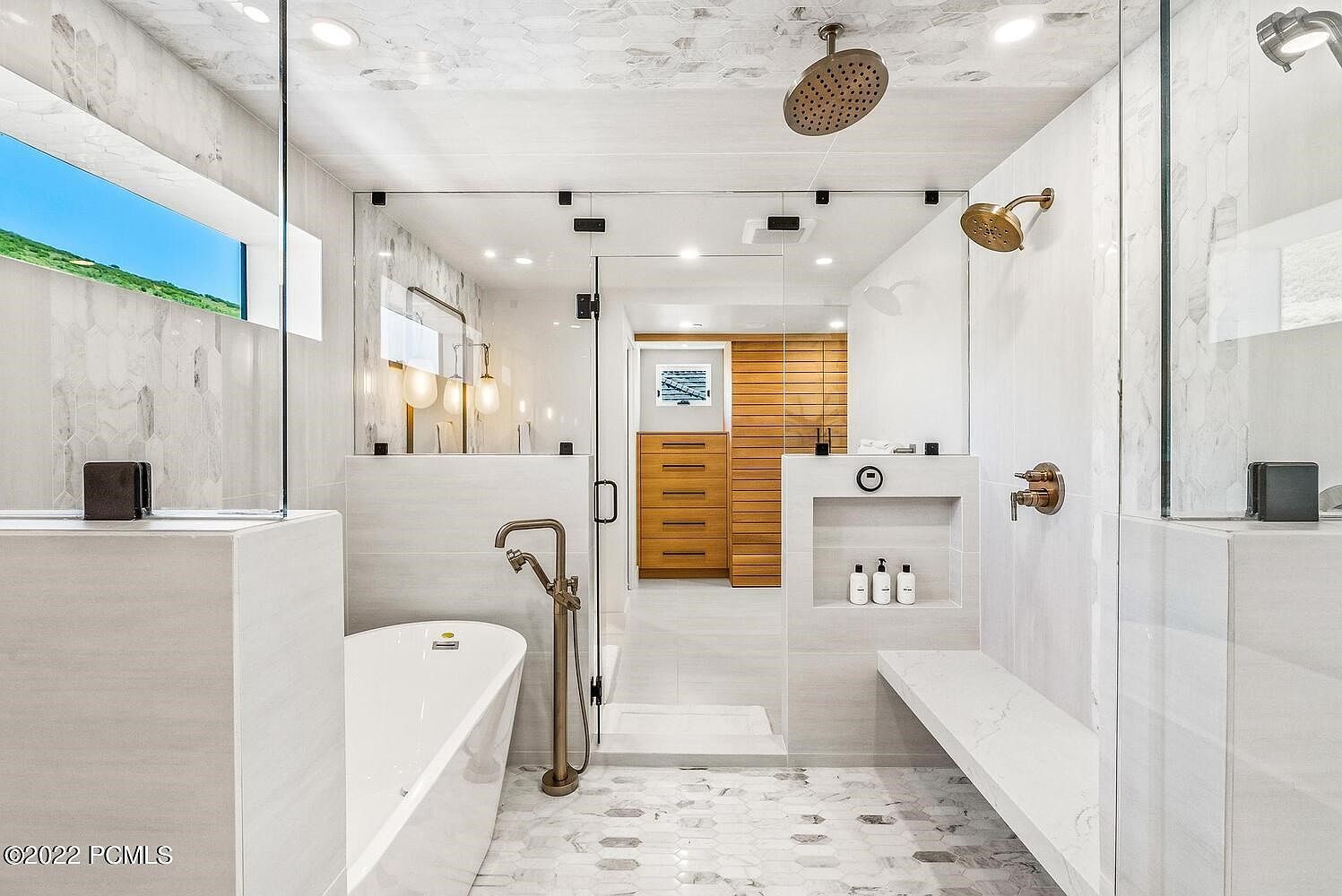 This is a luxurious primary bathroom featuring a freestanding bathtub with a modern faucet, a glass-enclosed shower with dual shower heads, and a built-in bench. The bathroom is adorned with white and gray marble tiles, creating a clean and elegant aesthetic. The perspective is from the entrance of the bathroom, showcasing the layout and high-end finishes.