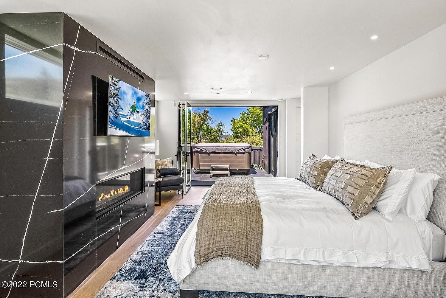 This is a luxurious primary bedroom featuring a modern fireplace with a TV mounted above it, a king-size bed with neutral-toned bedding, and an outdoor patio with a hot tub accessible through glass doors. The room is decorated in a contemporary style with clean lines and a neutral color palette, creating a serene and inviting atmosphere. The perspective is from the foot of the bed, looking towards the fireplace and patio.