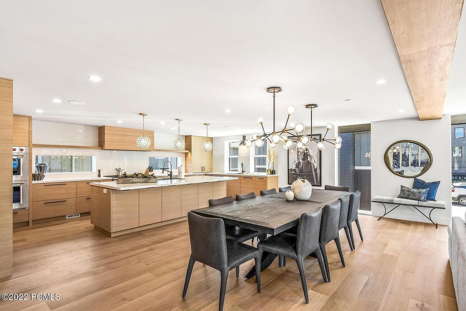 This interior shot showcases a modern dining area adjacent to a kitchen. The dining space features a long, dark wood table surrounded by gray upholstered chairs, illuminated by a contemporary chandelier. The kitchen area includes light wood cabinetry, a large island, and stainless steel appliances, creating a cohesive and stylish living space.
