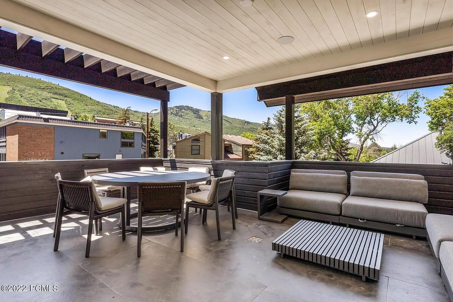 This image showcases a stylish outdoor patio or balcony area, featuring a dining set with a round table and chairs, along with a comfortable lounge area with a sofa and a modern coffee table. The space is partially covered, offering shade, and provides scenic views of the surrounding mountains and greenery, enhancing the property's appeal for outdoor living and entertaining.