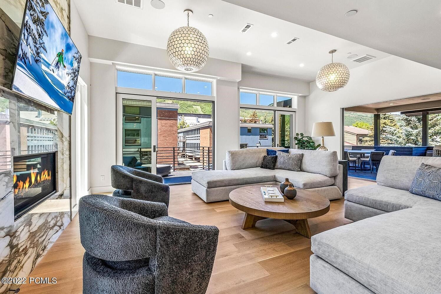 This is a bright and airy living room featuring hardwood floors, a modern fireplace with a TV above, and comfortable gray sectional sofas. Large windows and sliding glass doors provide ample natural light and access to an outdoor area. The room is decorated with stylish light fixtures and neutral tones, creating a welcoming and sophisticated atmosphere.