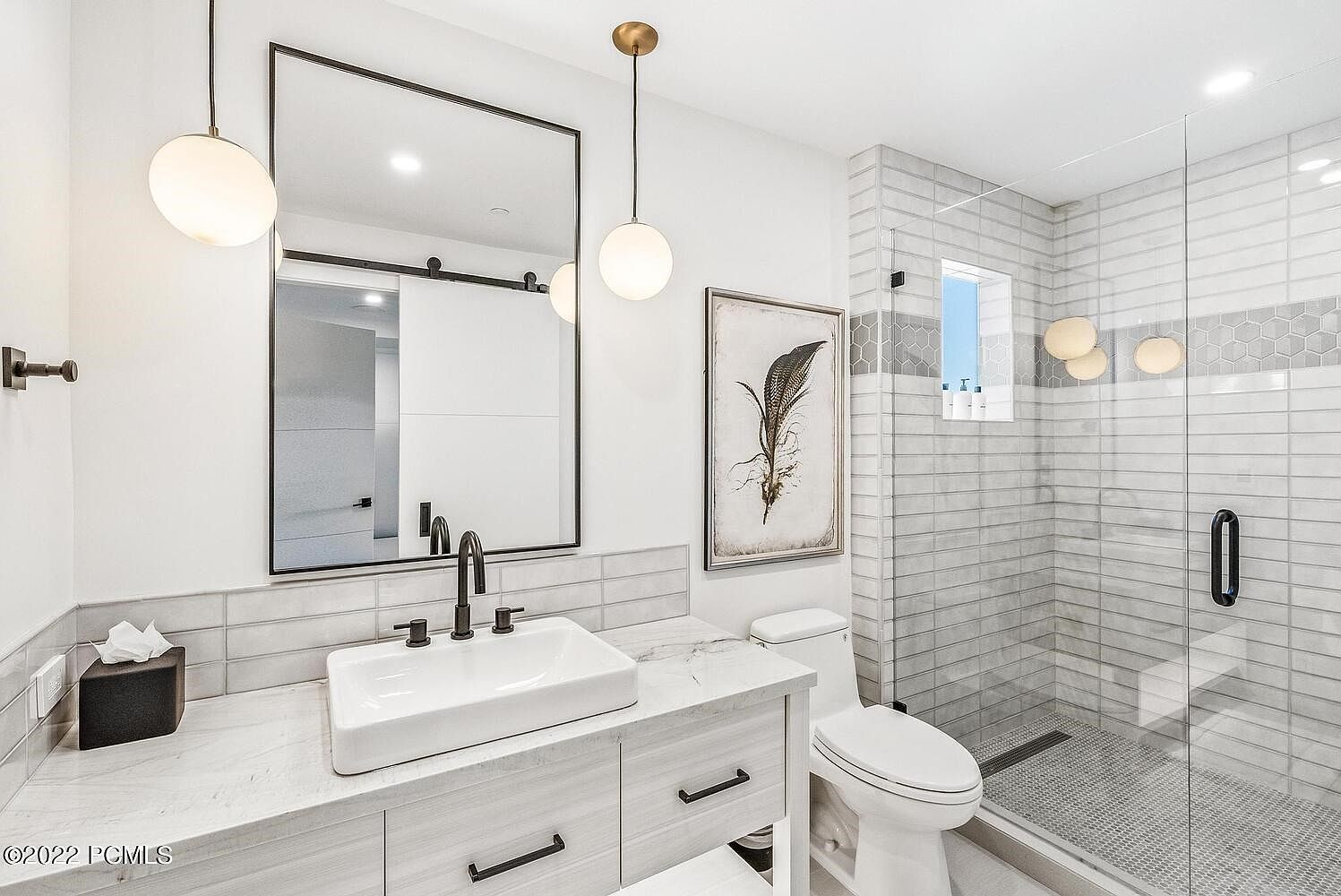 This is a well-lit primary bathroom featuring a modern design with a white vanity, a square vessel sink, and a black faucet. A large mirror with a black frame hangs above the vanity, reflecting a sliding door. The shower is enclosed with glass and features gray and white tiling, creating a clean and luxurious space.
