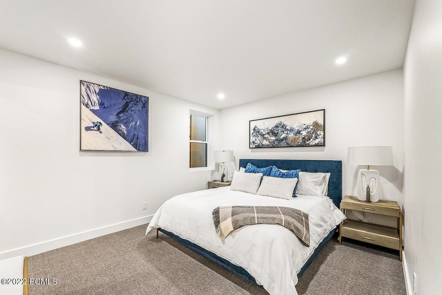 This is a well-lit bedroom featuring a blue upholstered bed with white linens and decorative pillows. Artwork adorns the white walls, and there are matching nightstands with lamps on either side of the bed. The room has a cozy and inviting atmosphere, perfect for a guest room.