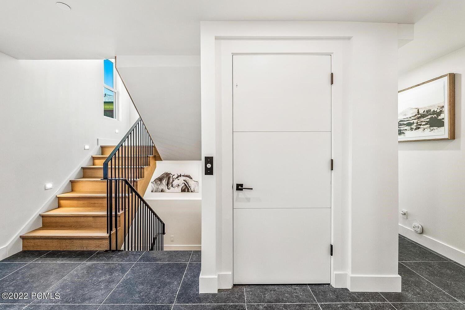 This interior shot showcases a modern hallway with a staircase. The staircase features wooden steps and a black metal railing, illuminated by recessed lighting. A white door with a sleek, minimalist design is prominent, and the flooring is dark tile, contributing to a contemporary aesthetic.