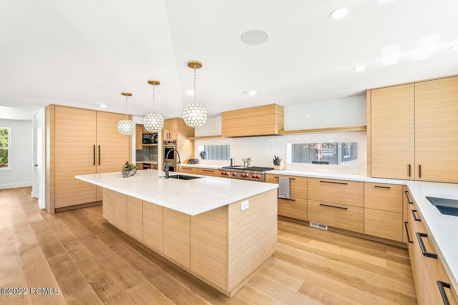 This is a bright and modern kitchen featuring light wood cabinetry and a large island with a white countertop. The kitchen is well-lit with pendant lighting and natural light from a window above the countertop. The hardwood floors add warmth to the space, creating an inviting atmosphere.