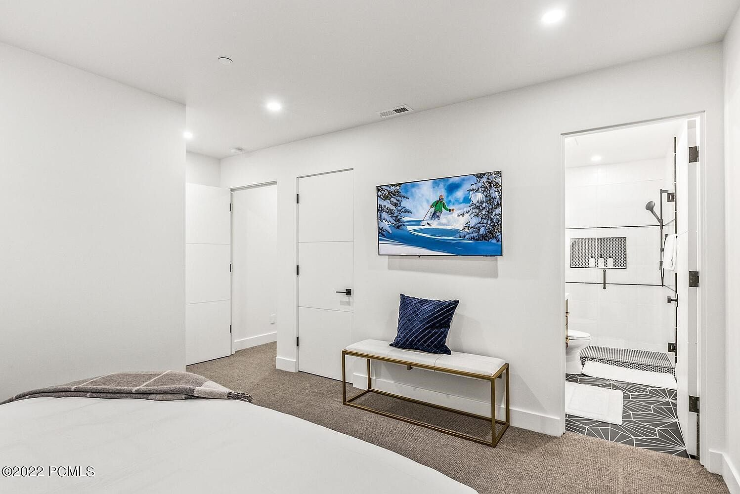 This is a bright and modern bedroom featuring a minimalist design with white walls and light-colored carpet. The room includes a bed, a bench with a blue pillow, and a wall-mounted television displaying a winter scene. An open doorway leads to a contemporary bathroom with black and white patterned tile, enhancing the room's appeal for potential buyers.