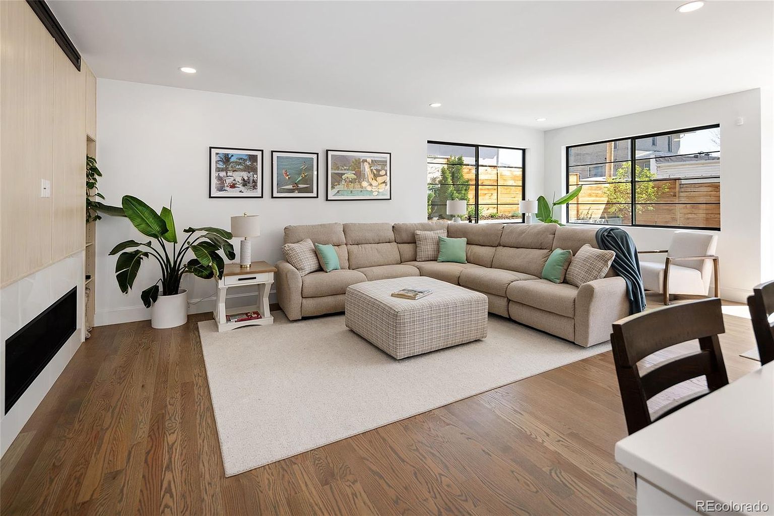 This is a bright and inviting living room featuring hardwood floors and a large sectional sofa with decorative pillows. Three framed art pieces hang above the sofa, and large windows provide ample natural light. An area rug defines the seating area, and a patterned ottoman serves as a coffee table.