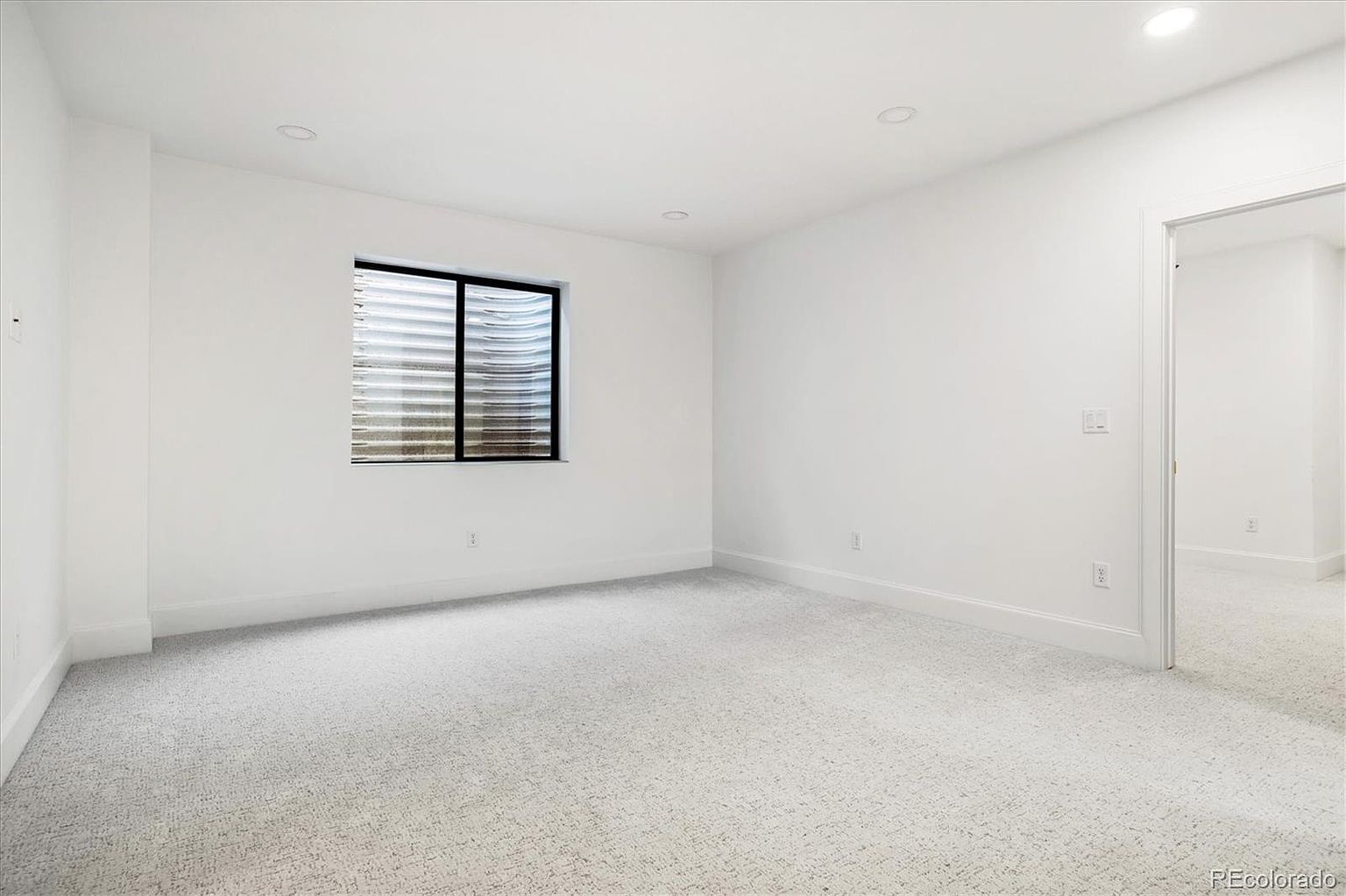 This is a bright, minimalist bedroom with white walls, trim, and carpet. A single window with horizontal blinds provides natural light. The room appears spacious and clean, ready for personalization.