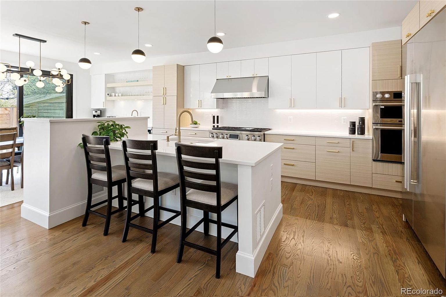 This is a bright and modern kitchen featuring a large island with seating, white and light wood cabinetry, and stainless steel appliances. The kitchen is well-lit with pendant lighting and recessed lights, highlighting the clean lines and contemporary design. The hardwood flooring adds warmth to the space.