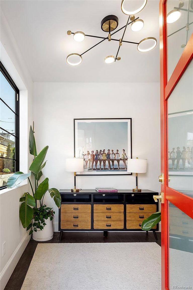 This interior shot showcases a stylish hallway featuring a modern chandelier, a console table with lamps and artwork, and a large potted plant. The space is bright and airy, with a clean, contemporary aesthetic. A red door adds a pop of color and visual interest.