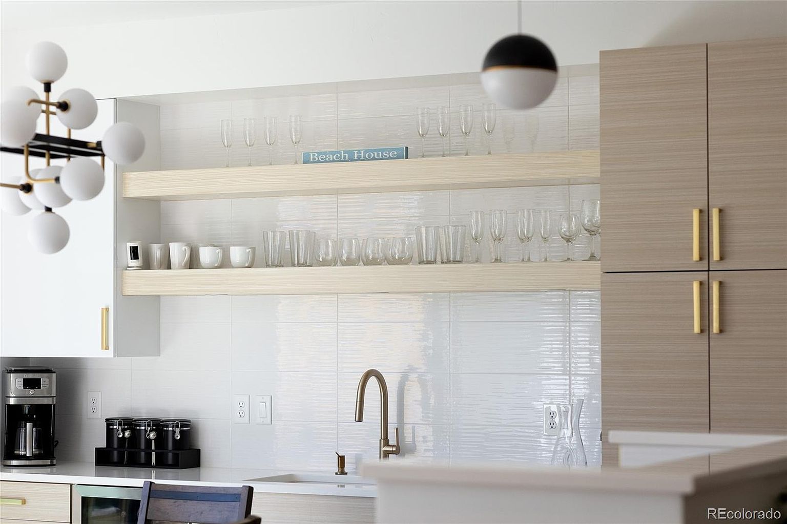 This is a bright and modern kitchen featuring white and light wood cabinetry with gold hardware. Open shelving displays glassware and decorative items, complemented by a stylish light fixture. The kitchen is well-lit and appears clean and organized, creating an inviting atmosphere.