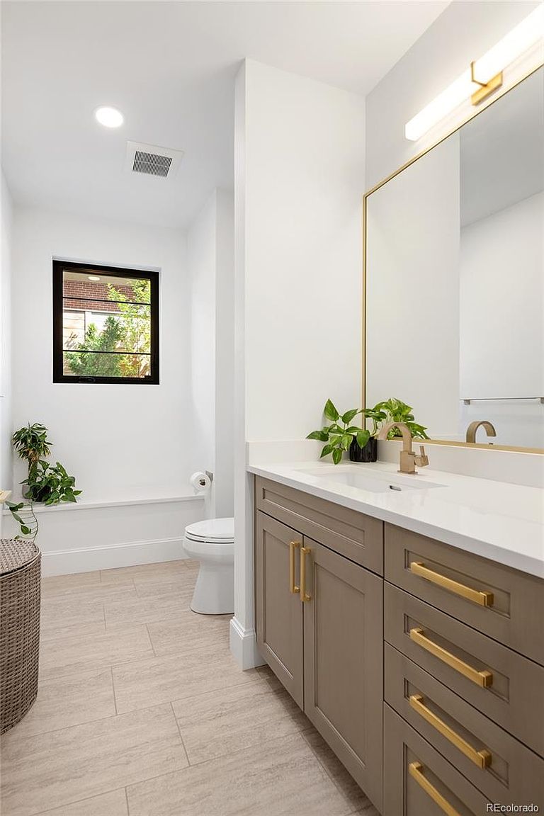 This is a well-lit bathroom featuring a modern vanity with taupe-colored cabinets and gold hardware, topped with a white countertop and a gold-framed mirror. A toilet is visible to the left, and the room is finished with light-colored tile flooring. The overall impression is clean, contemporary, and stylish.