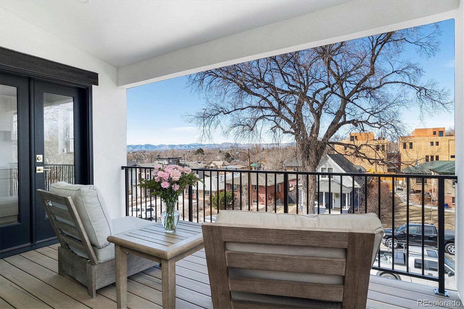 This image showcases a charming balcony with comfortable outdoor seating, including two cushioned chairs and a small wooden table adorned with a vase of pink flowers. The balcony features a black metal railing, offering an unobstructed view of the surrounding neighborhood, distant mountains, and a large tree. The overall impression is one of relaxation and tranquility.