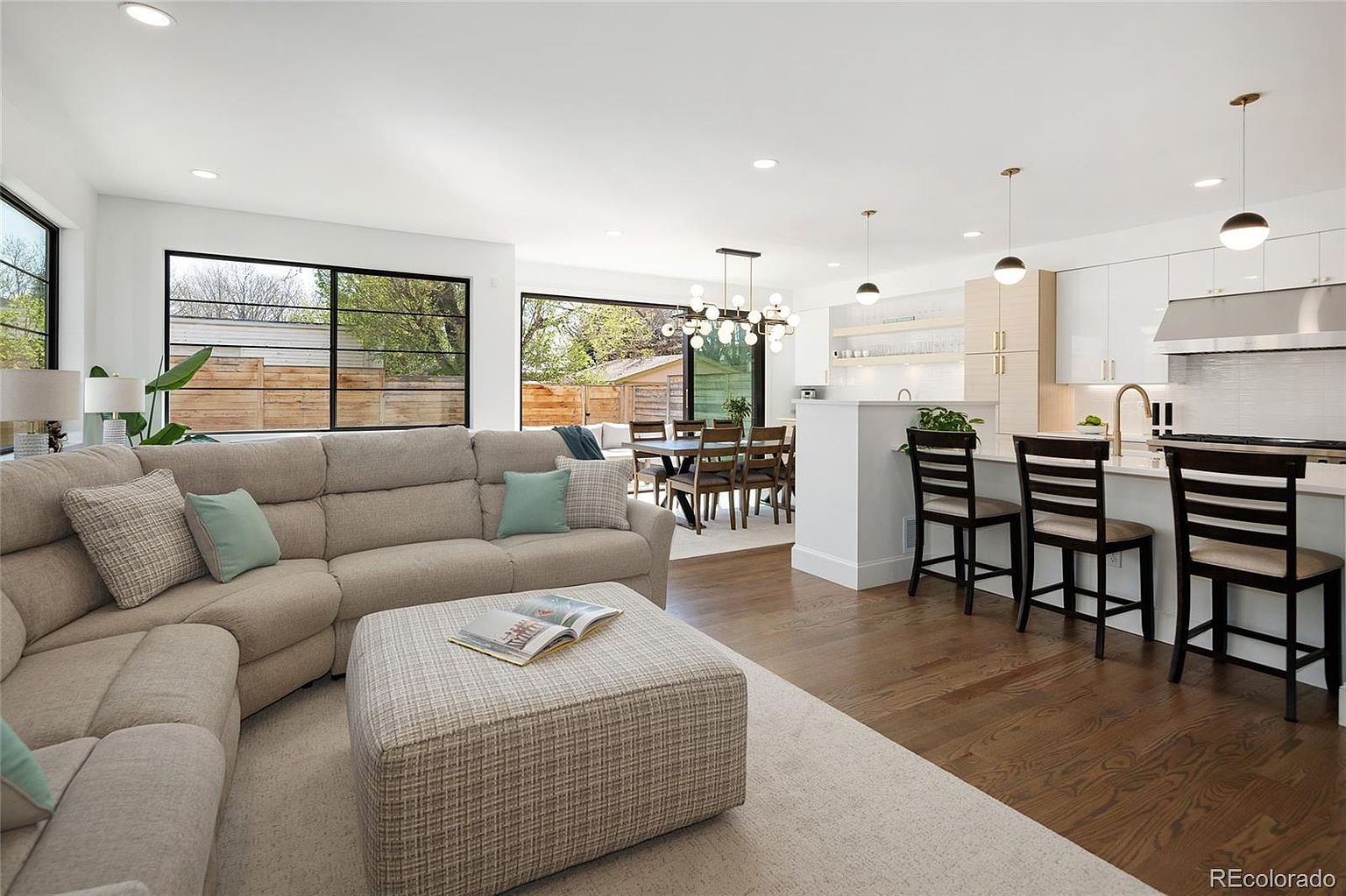 This is an interior shot of a modern living room that seamlessly blends into the dining and kitchen areas. The living room features a large, comfortable sectional sofa with throw pillows, an ottoman, and a neutral-toned rug. Large windows provide ample natural light and views of the outdoors, while the open floor plan creates a spacious and inviting atmosphere.
