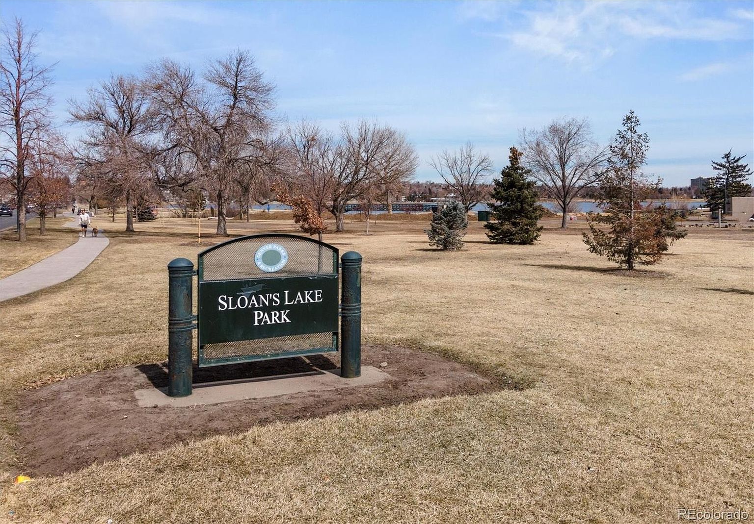This image showcases Sloan's Lake Park, featuring a prominent park sign in the foreground. The park's landscape includes a mix of grassy areas and bare trees, suggesting a transition season. In the background, a glimpse of the lake and distant buildings adds depth to the scene, creating a serene and inviting atmosphere.