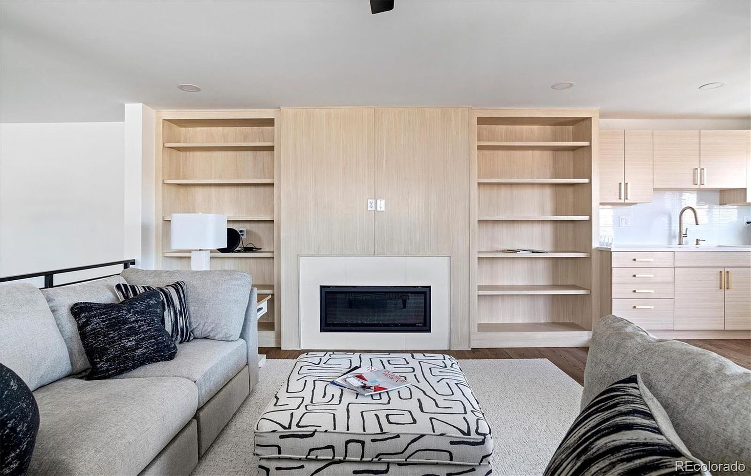 This is an interior shot of a living room featuring a modern fireplace with built-in shelving on either side. The room is furnished with a comfortable gray sofa adorned with decorative pillows and a patterned ottoman. The overall aesthetic is clean, bright, and inviting, suggesting a cozy and stylish living space.