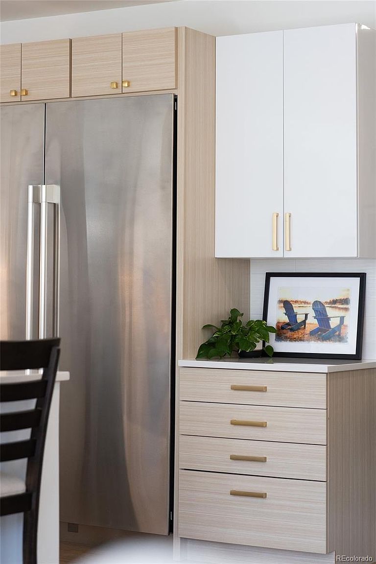 This image showcases a modern kitchen detail, focusing on the cabinetry and appliances. A stainless steel refrigerator is flanked by light wood-grain cabinets and drawers with sleek, gold hardware. A framed picture and a small plant add a touch of personality to the countertop.