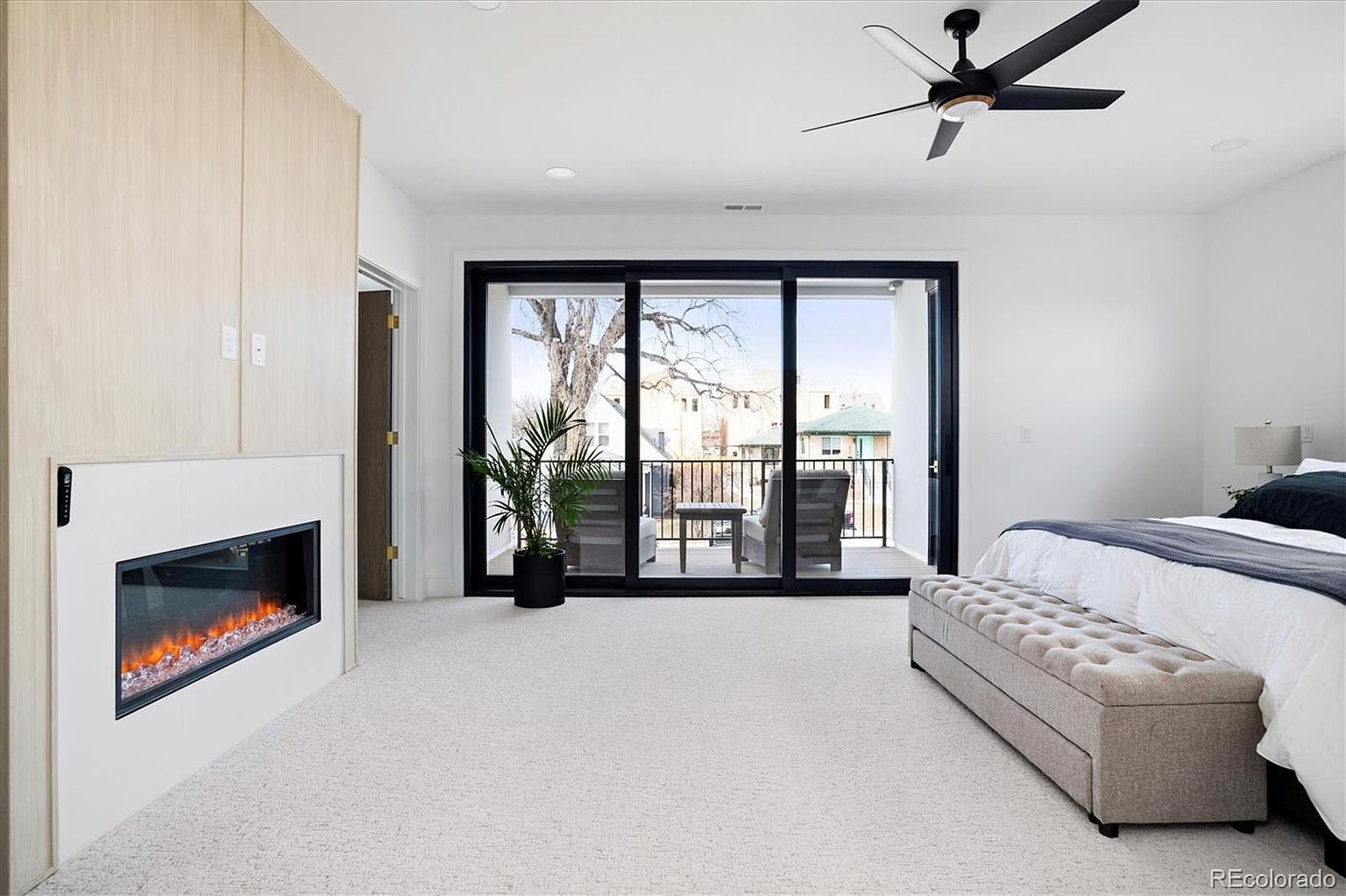 This is a bright and airy primary bedroom featuring a modern fireplace, a large sliding glass door leading to a balcony, and a neutral color palette. The room is carpeted and includes a bed with a tufted bench at the foot. The overall impression is clean, contemporary, and inviting.