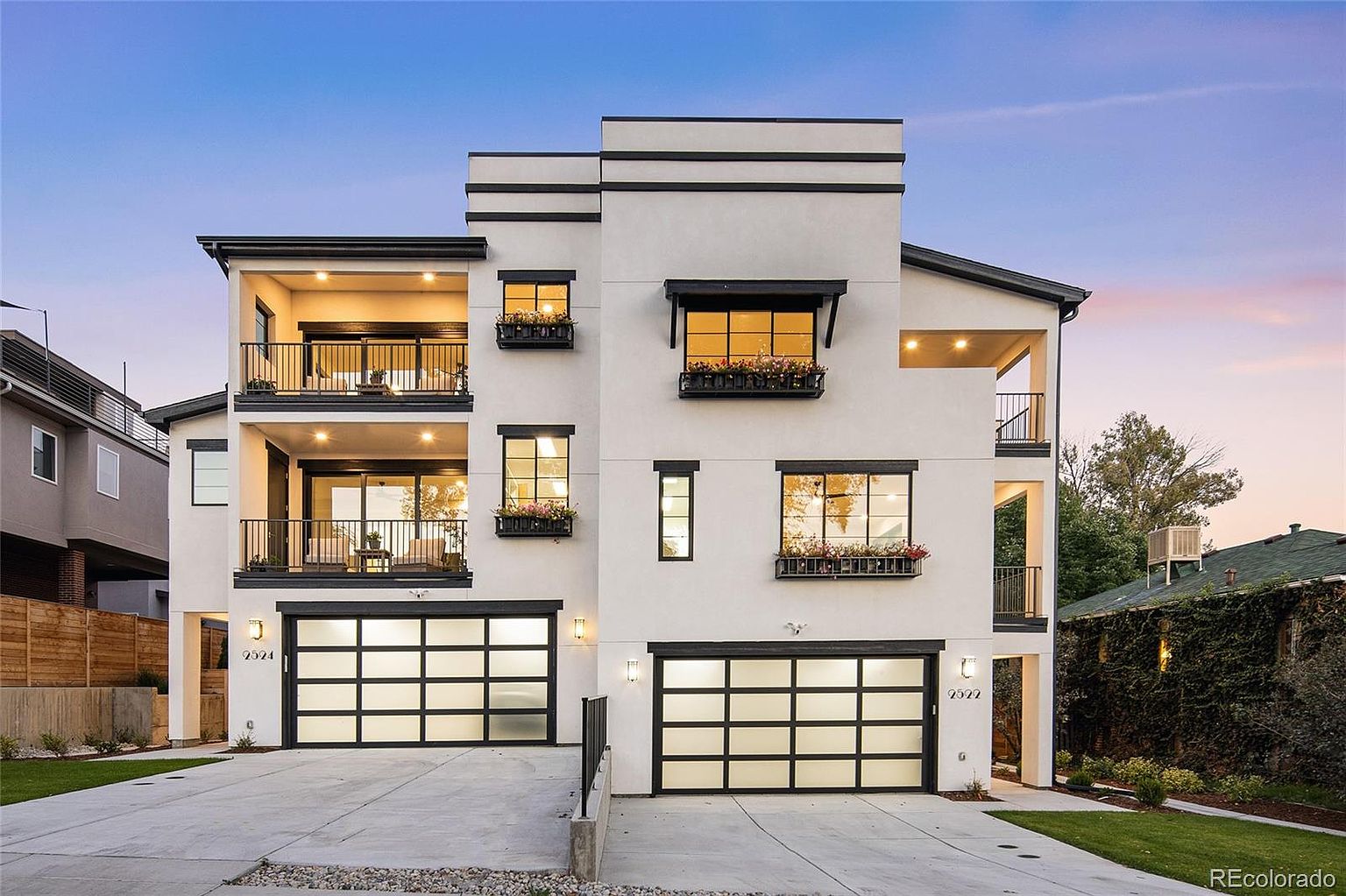 This is a front view of a modern, multi-story home featuring a white facade with black trim. The house has two garage doors, multiple balconies with black railings, and window boxes filled with flowers. The overall impression is stylish and contemporary, with a clean and sophisticated aesthetic.