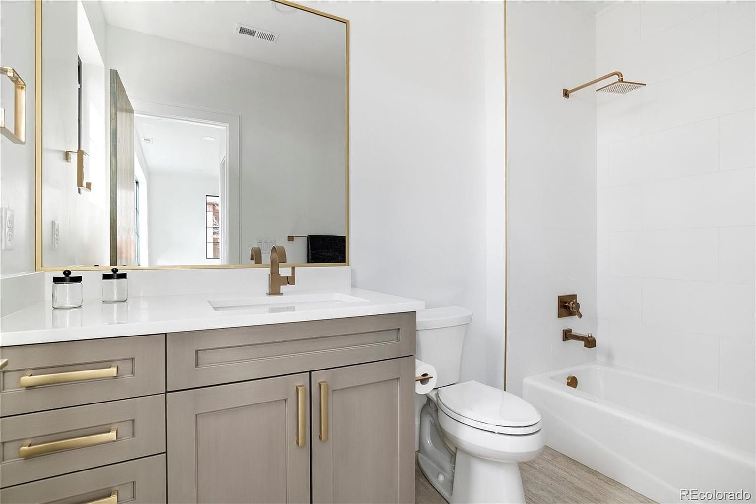 This is a well-lit bathroom featuring a vanity with a gray cabinet and white countertop, complemented by gold hardware and a matching framed mirror. A white toilet sits beside the vanity, and a bathtub with a showerhead is visible in the background, all contributing to a clean and modern aesthetic. The perspective is from the doorway, showcasing the entire bathroom.