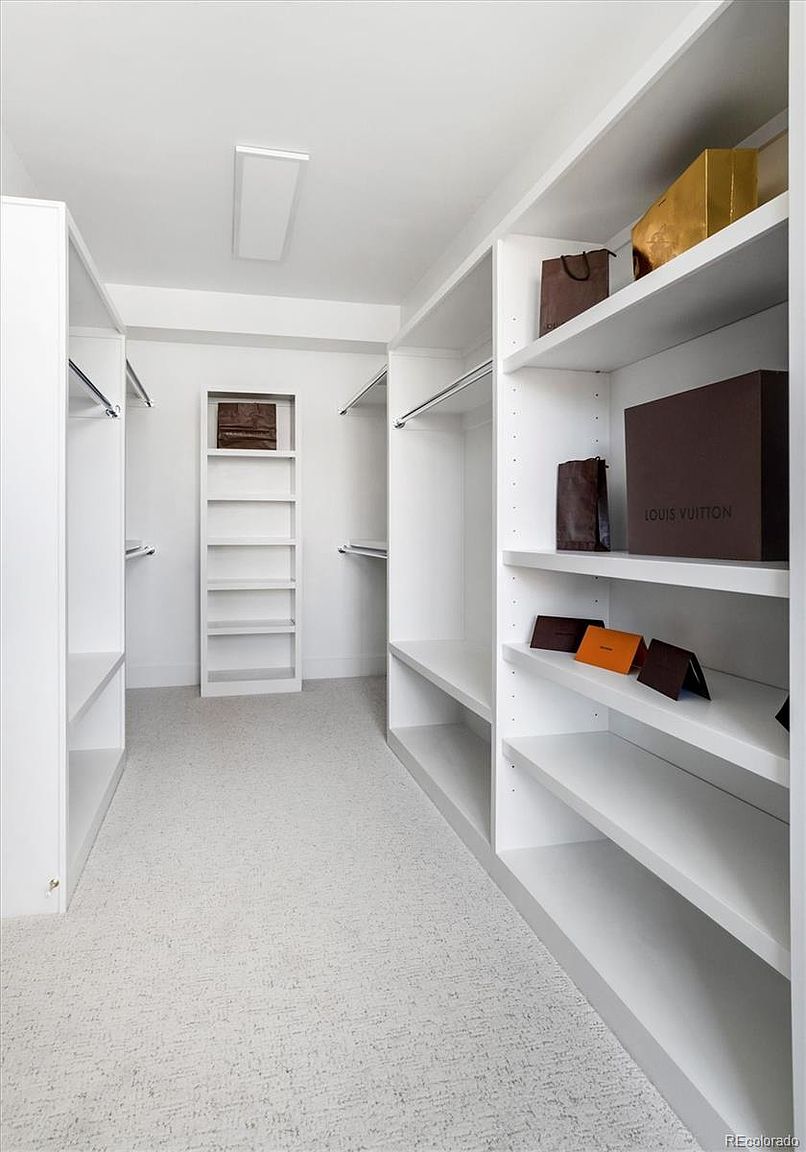 This is a well-organized walk-in closet featuring white shelving and hanging rods. The closet is spacious and brightly lit, with a neutral-colored carpet. Several shelves display designer shopping bags and accessories, suggesting a luxurious and organized space.