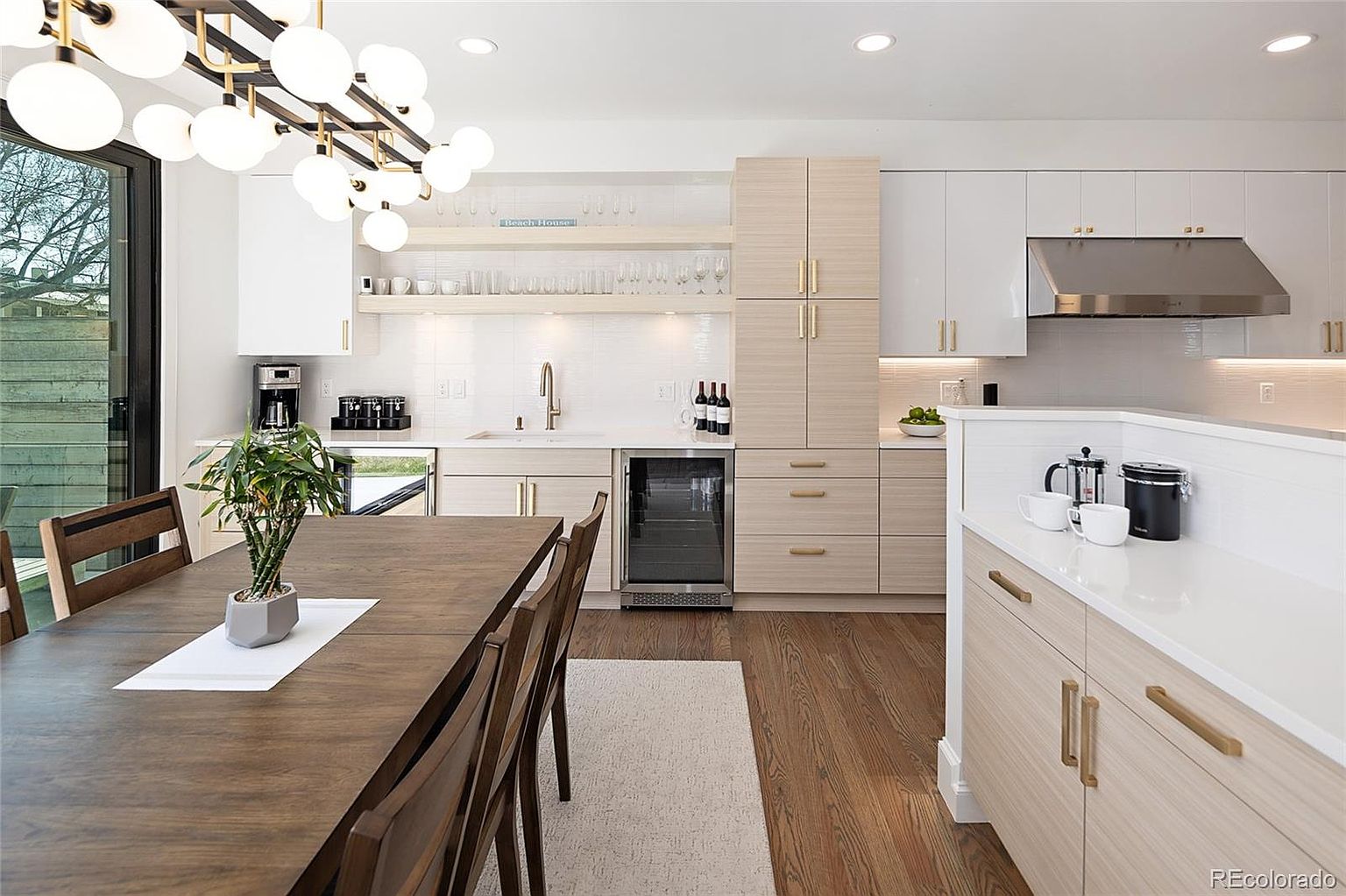 This is a bright and modern kitchen featuring a dining area. The kitchen boasts sleek cabinetry with gold hardware, stainless steel appliances, and a wine fridge. A large dining table with wooden chairs sits adjacent to the kitchen area, illuminated by a modern chandelier, creating an inviting and functional space.