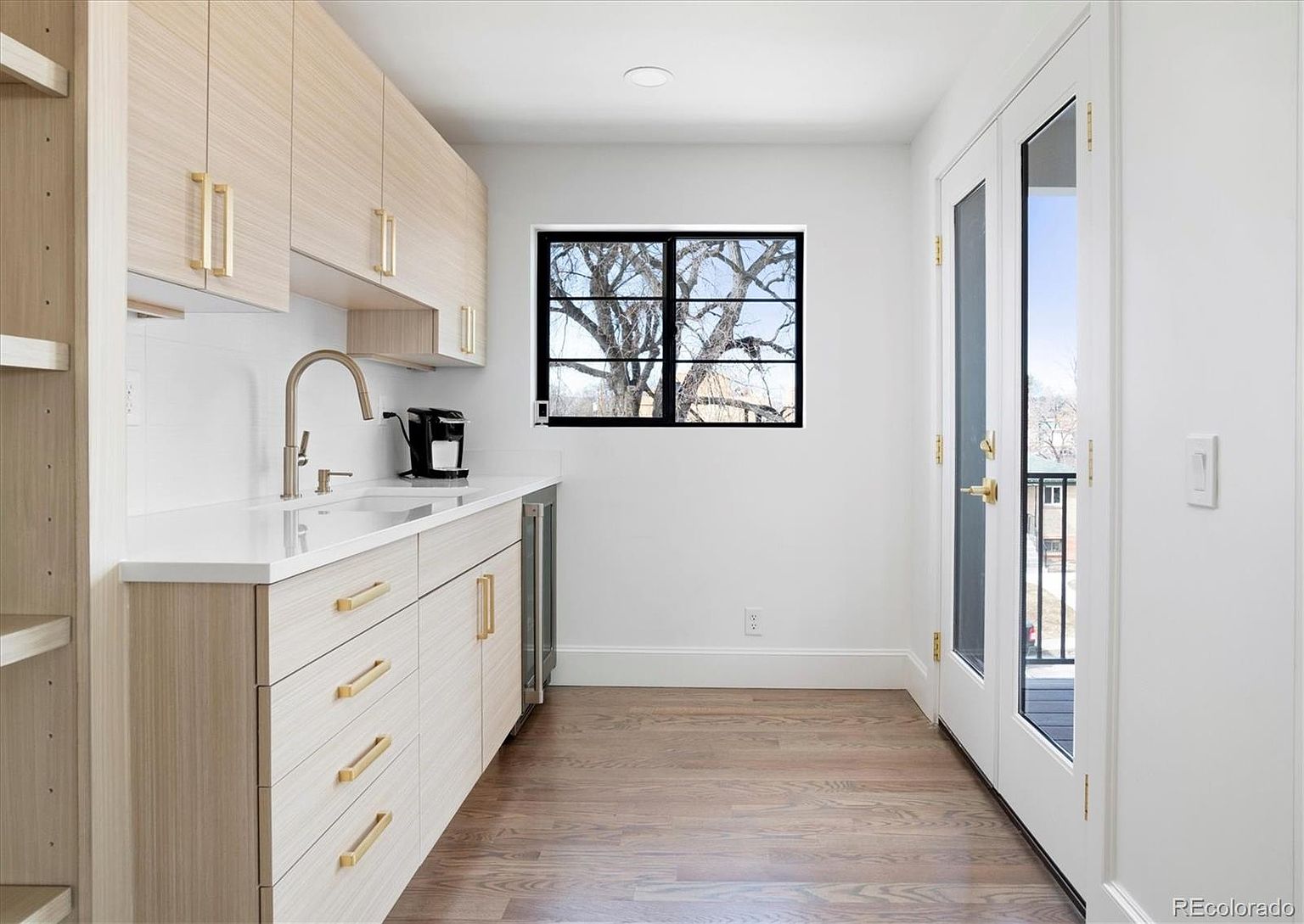 This interior shot showcases a modern kitchen area with light wood cabinetry and gold hardware, complemented by a white countertop and backsplash. A stainless steel faucet and a small appliance sit on the counter, while a window provides natural light and a view of a tree. Double doors lead to a balcony, enhancing the space's appeal.