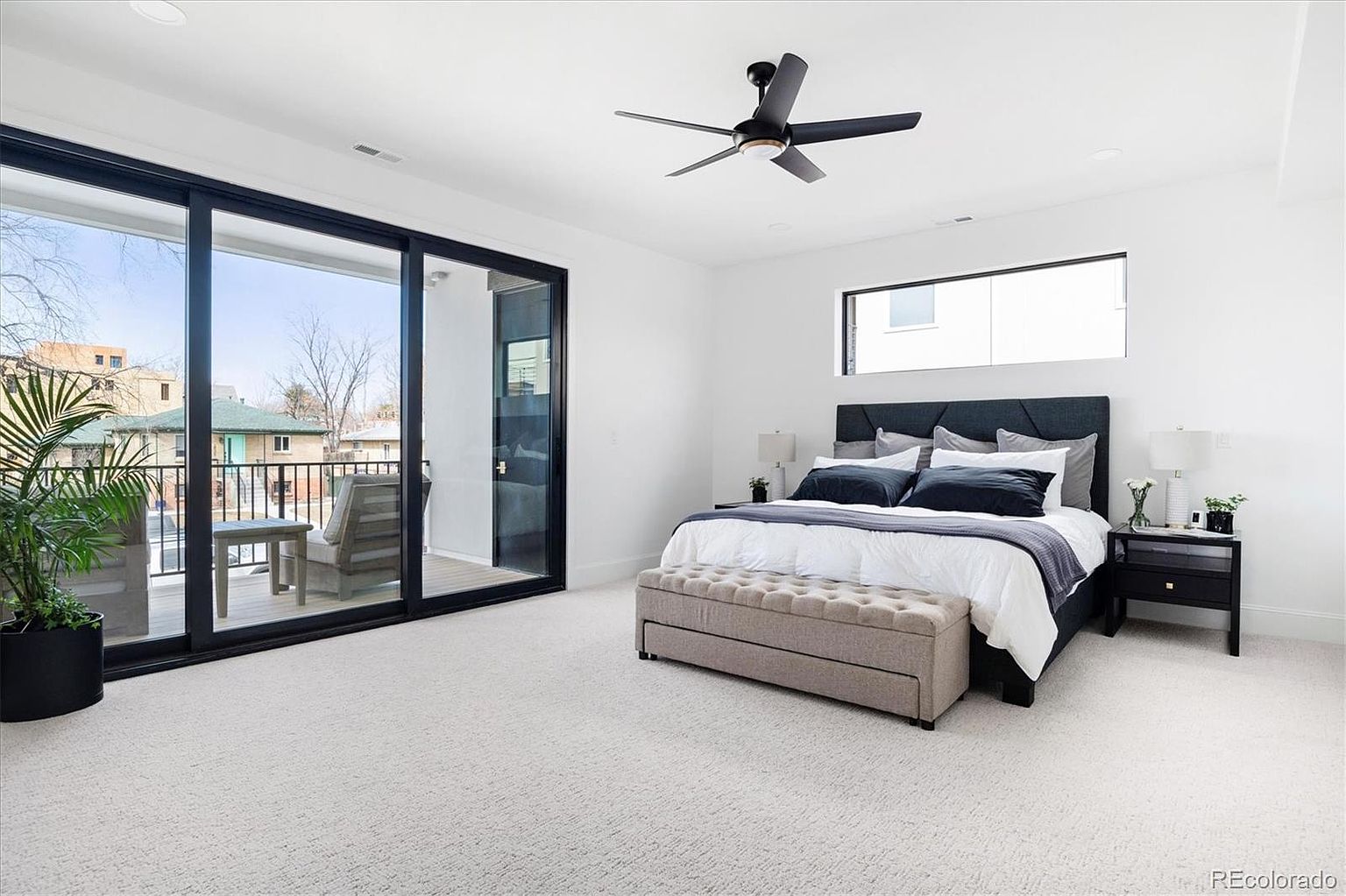 This is a bright and airy primary bedroom featuring a large sliding glass door leading to a balcony, allowing for ample natural light. The room is decorated in a modern style with a neutral color palette, including a gray upholstered headboard, white bedding with gray accents, and a beige ottoman at the foot of the bed. A black ceiling fan and a small window above the bed add to the room's contemporary feel.
