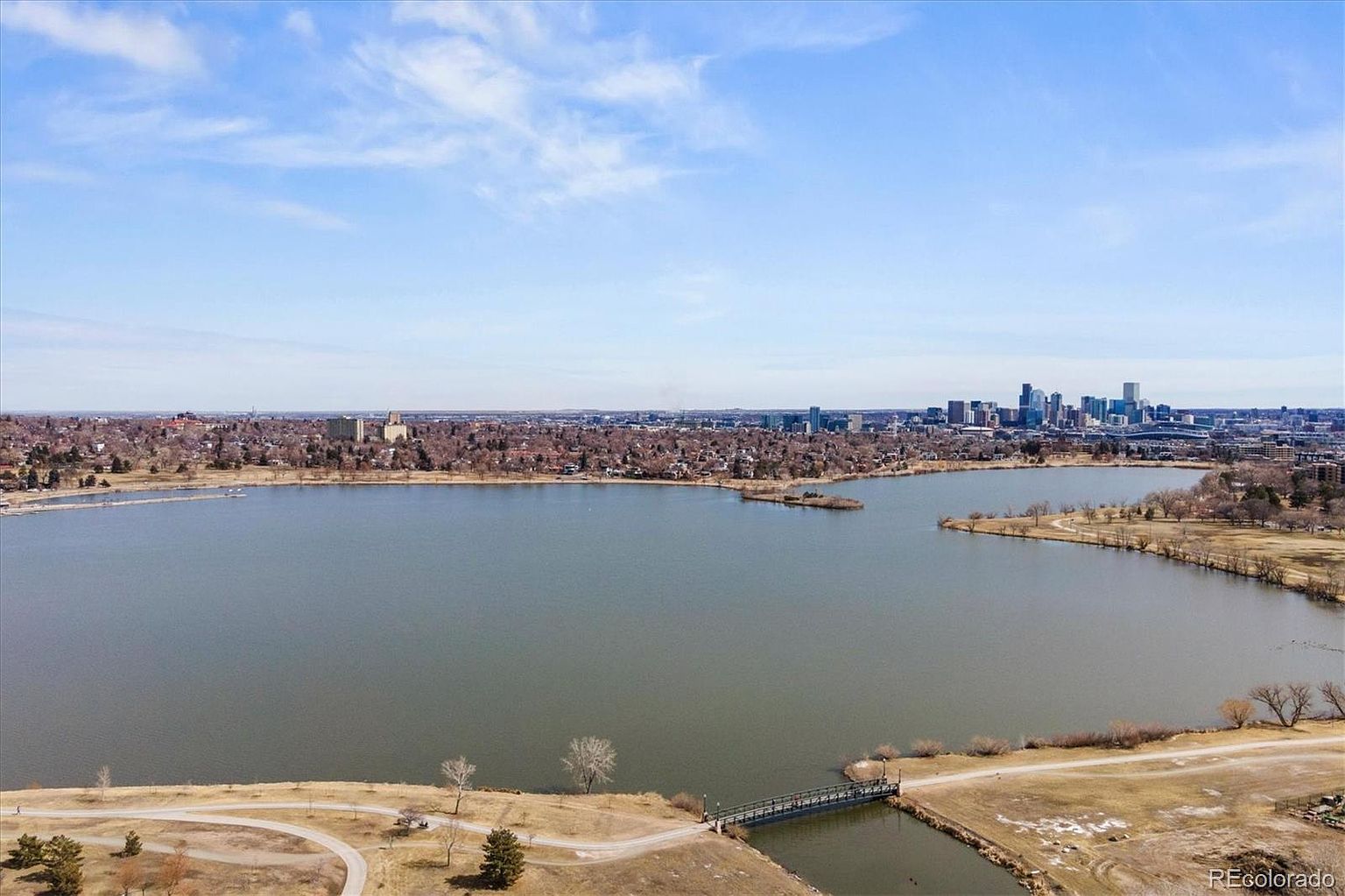 This aerial view showcases a large lake surrounded by residential areas and a distant city skyline. A small bridge connects a grassy area to the mainland, and the water appears calm and expansive. The image provides a sense of the property's proximity to both nature and urban amenities.