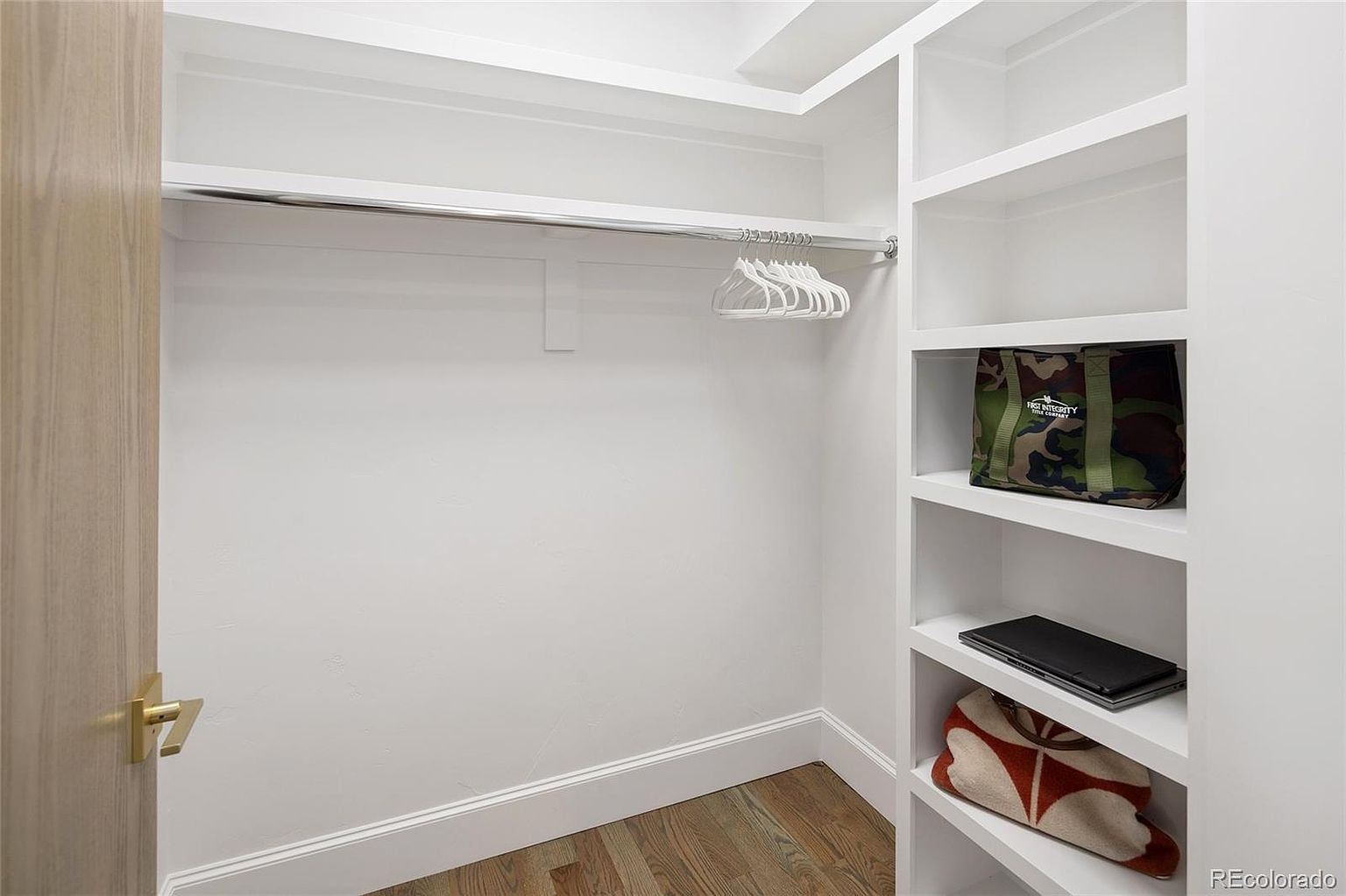 This is a well-organized walk-in closet featuring a chrome clothing rod with white hangers and built-in white shelving. The shelves hold items such as a camouflage tote bag, a laptop, and a patterned blanket. The closet has a clean, modern aesthetic with light wood flooring and white trim.