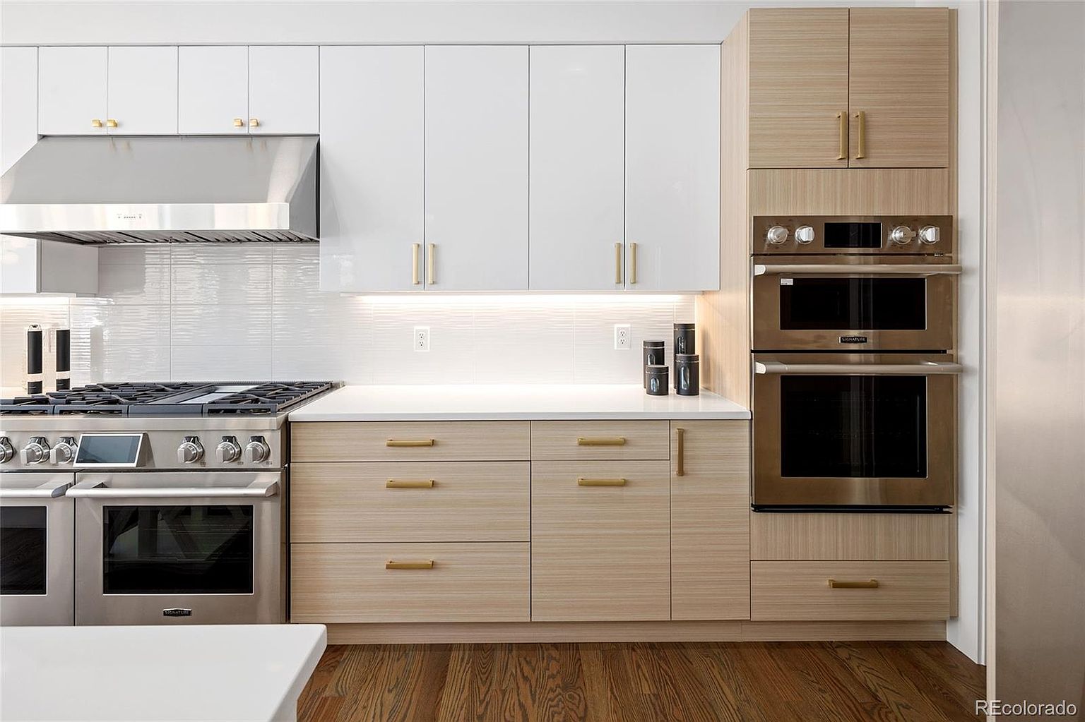 A well-lit kitchen showcases a modern design with a combination of white and light wood-grain cabinetry, complemented by sleek gold hardware. Stainless steel appliances, including a range and double oven, add a touch of luxury. The hardwood flooring and white countertops contribute to the clean and contemporary aesthetic.