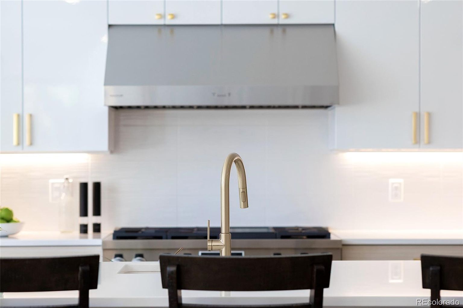 This is a modern kitchen featuring white cabinets with gold hardware and a stainless steel range hood. A gold faucet is centered in the frame, and dark wood chairs are partially visible in the foreground. The overall impression is clean and stylish.