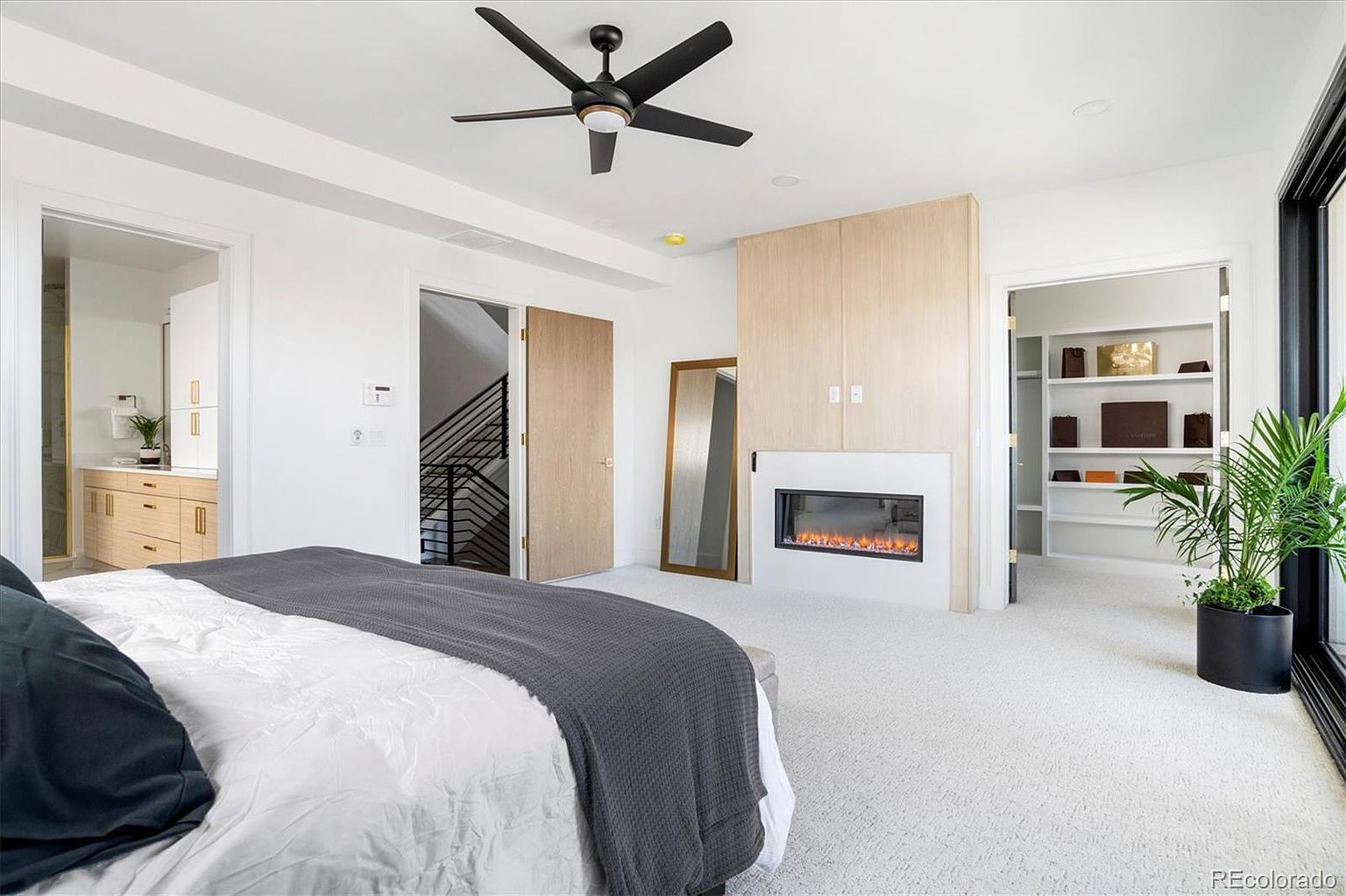 This is a bright and spacious primary bedroom featuring a modern fireplace, a ceiling fan, and a walk-in closet. The room is decorated in a minimalist style with white walls and light-colored wood accents. The bed is neatly made with a gray throw blanket, and a potted plant adds a touch of greenery.