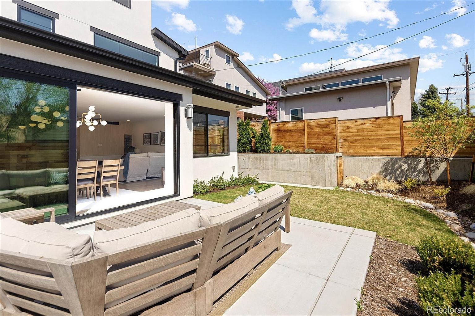 This image showcases the rear exterior of a modern home, highlighting a well-manicured backyard and patio area. A large sliding glass door provides seamless indoor-outdoor living, leading to a patio furnished with a stylish outdoor sofa. The backyard features a combination of lawn, garden beds, and a wooden fence, creating a private and inviting outdoor space.
