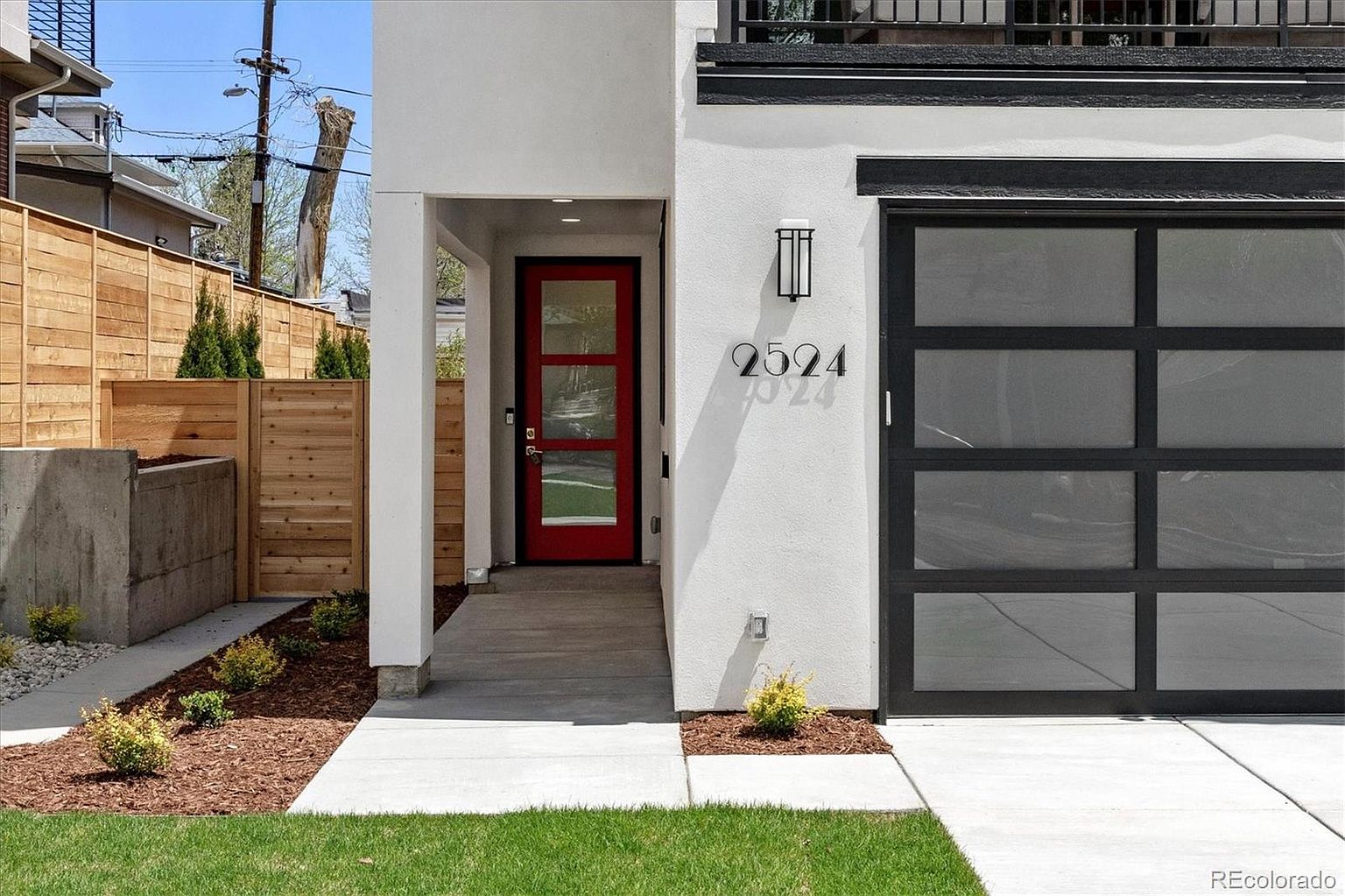 This image showcases the modern entryway of a home, featuring a striking red front door with glass panels, flanked by white stucco walls. The house number '2524' is prominently displayed, and a contemporary light fixture adds to the aesthetic. A sleek, black-framed garage door is visible to the right, and the landscaping includes a small patch of green lawn and mulch beds with young plants, creating a welcoming and stylish curb appeal.