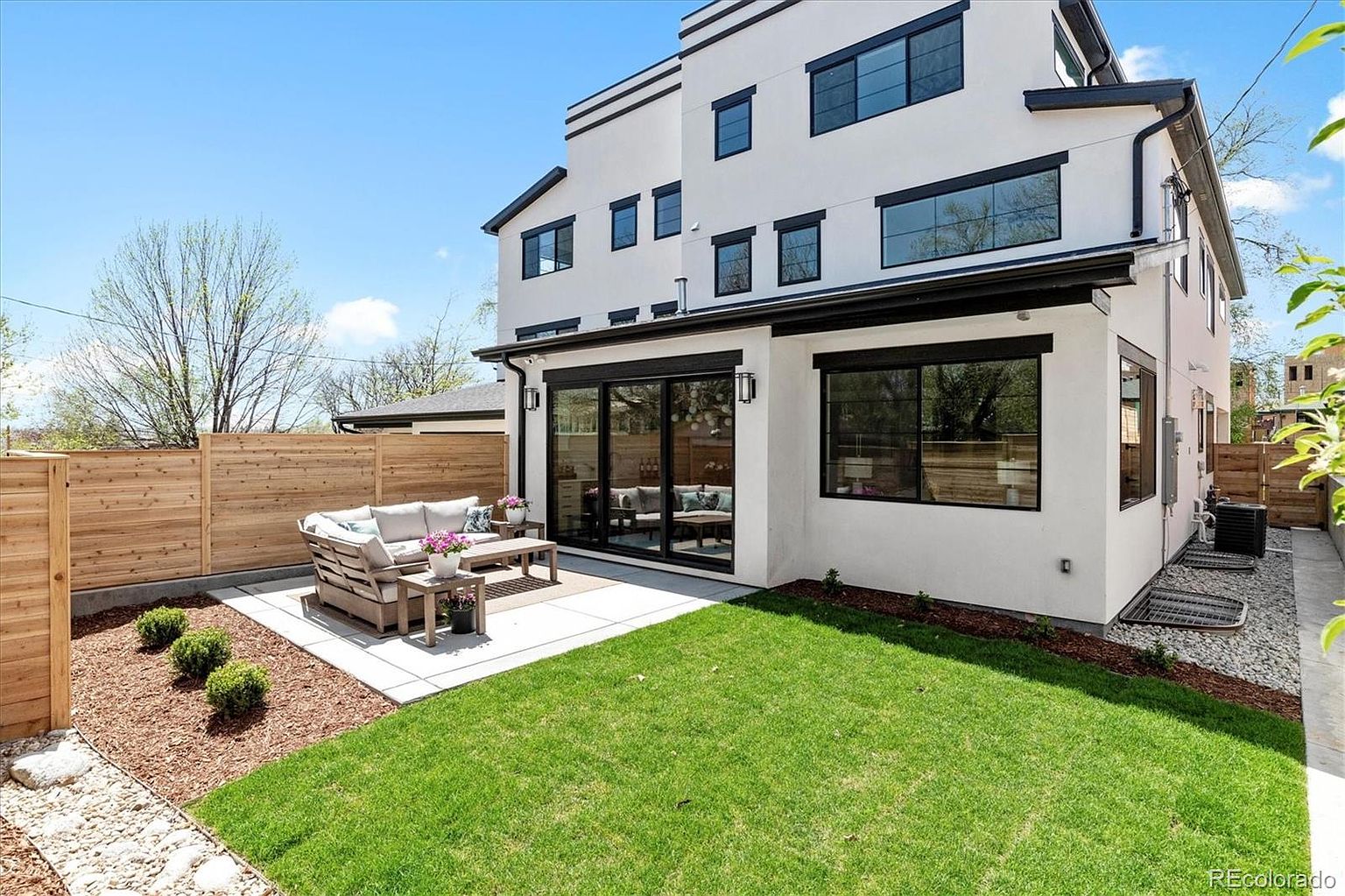 This image showcases a well-maintained backyard featuring a modern patio set on a concrete slab, surrounded by lush green grass and neatly arranged landscaping. A contemporary white house with black-framed windows provides a stylish backdrop, complemented by a wooden privacy fence. The scene evokes a sense of tranquility and outdoor living.